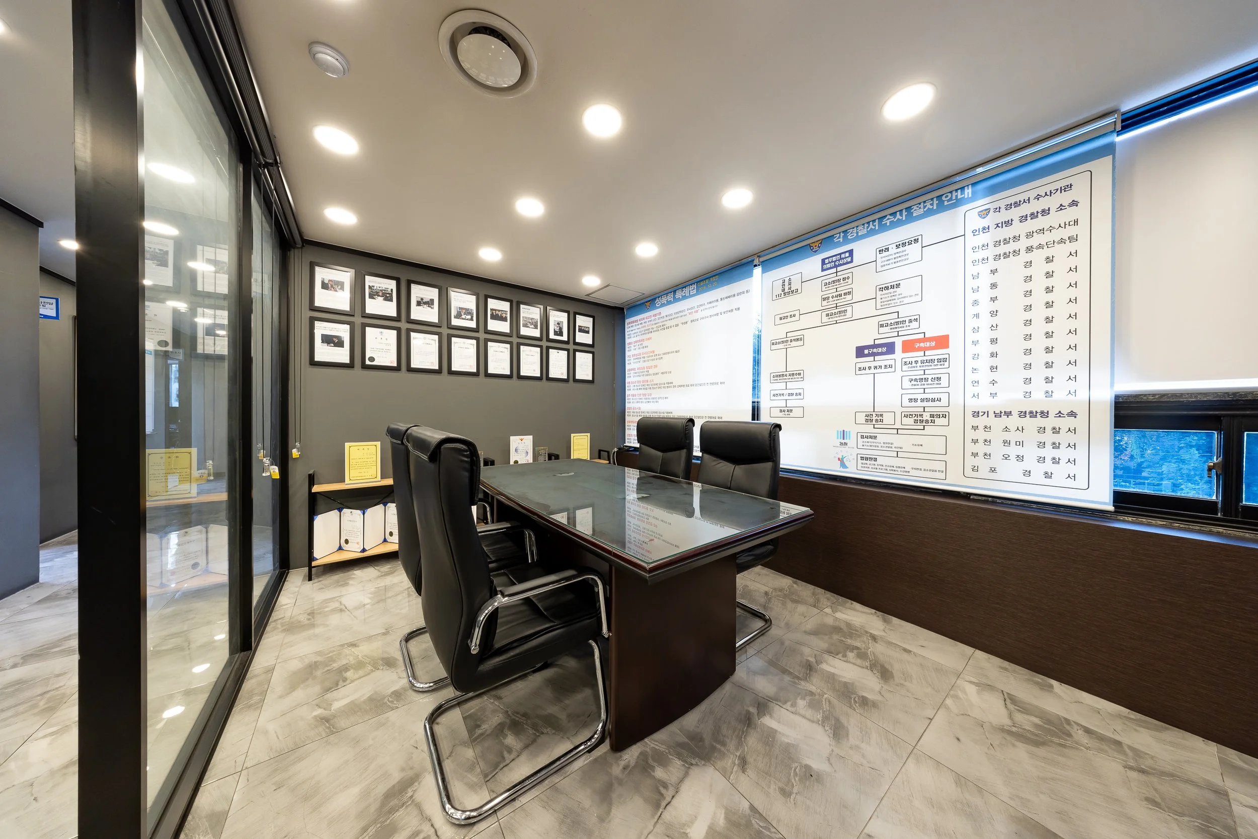 An office conference room with a glass-top table, black leather chairs, framed certificates on the wall, and informational posters on the side wall.
