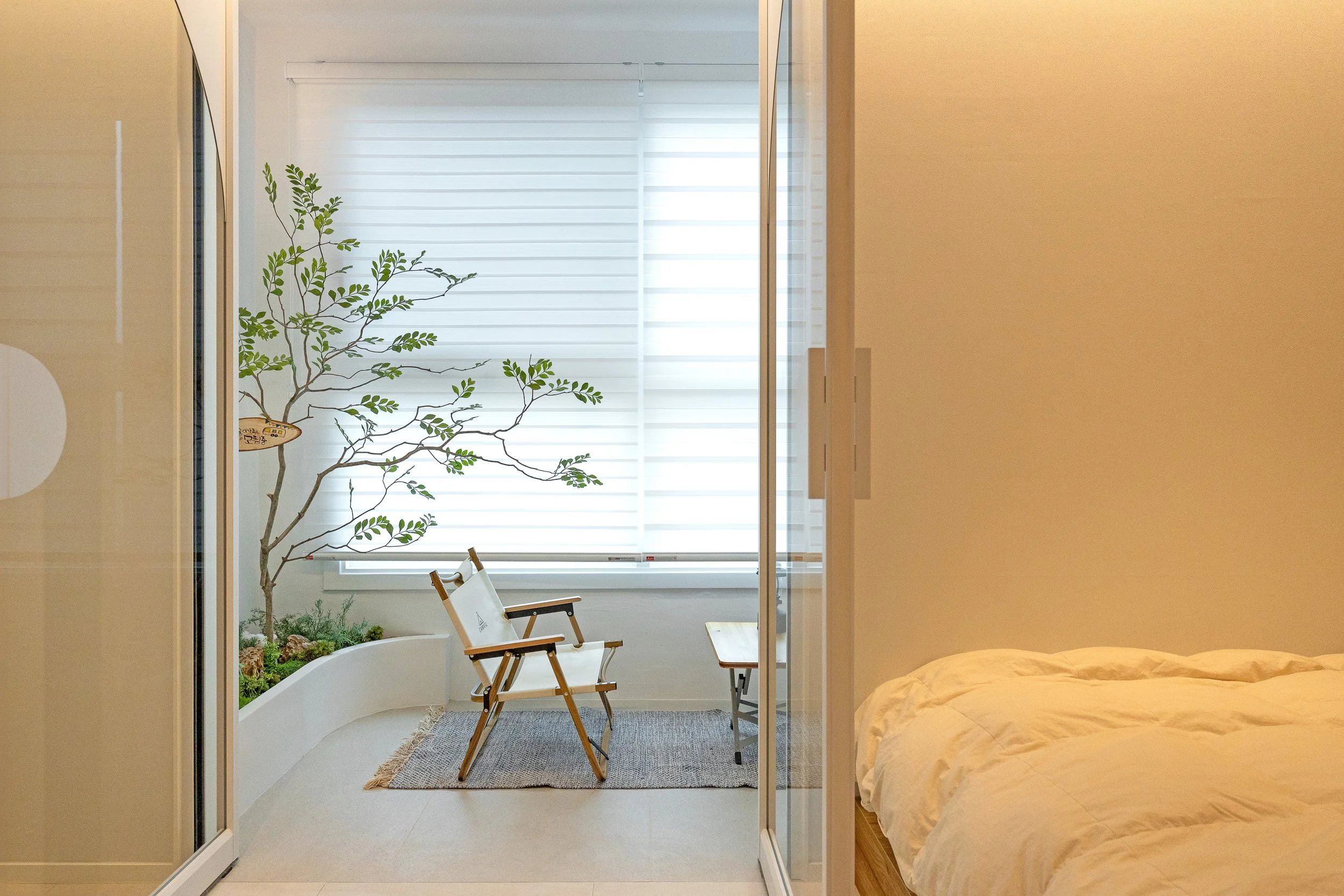 A cozy bedroom with a view into an enclosed balcony area. The balcony has a small tree, two chairs, a table, a rectangular rug, and white blinds on the window. The bedroom has a bed with white bedding.