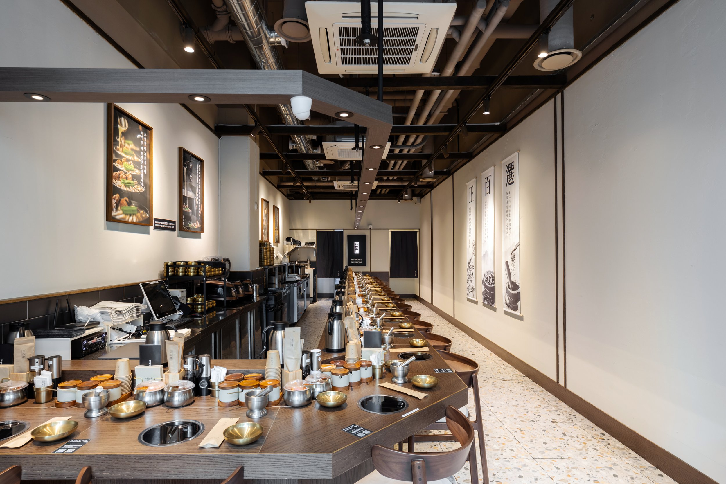 Interior of a modern sushi restaurant with a long bar table set with bowls, cups, and utensils, and a wall with framed pictures and Japanese calligraphy scrolls.