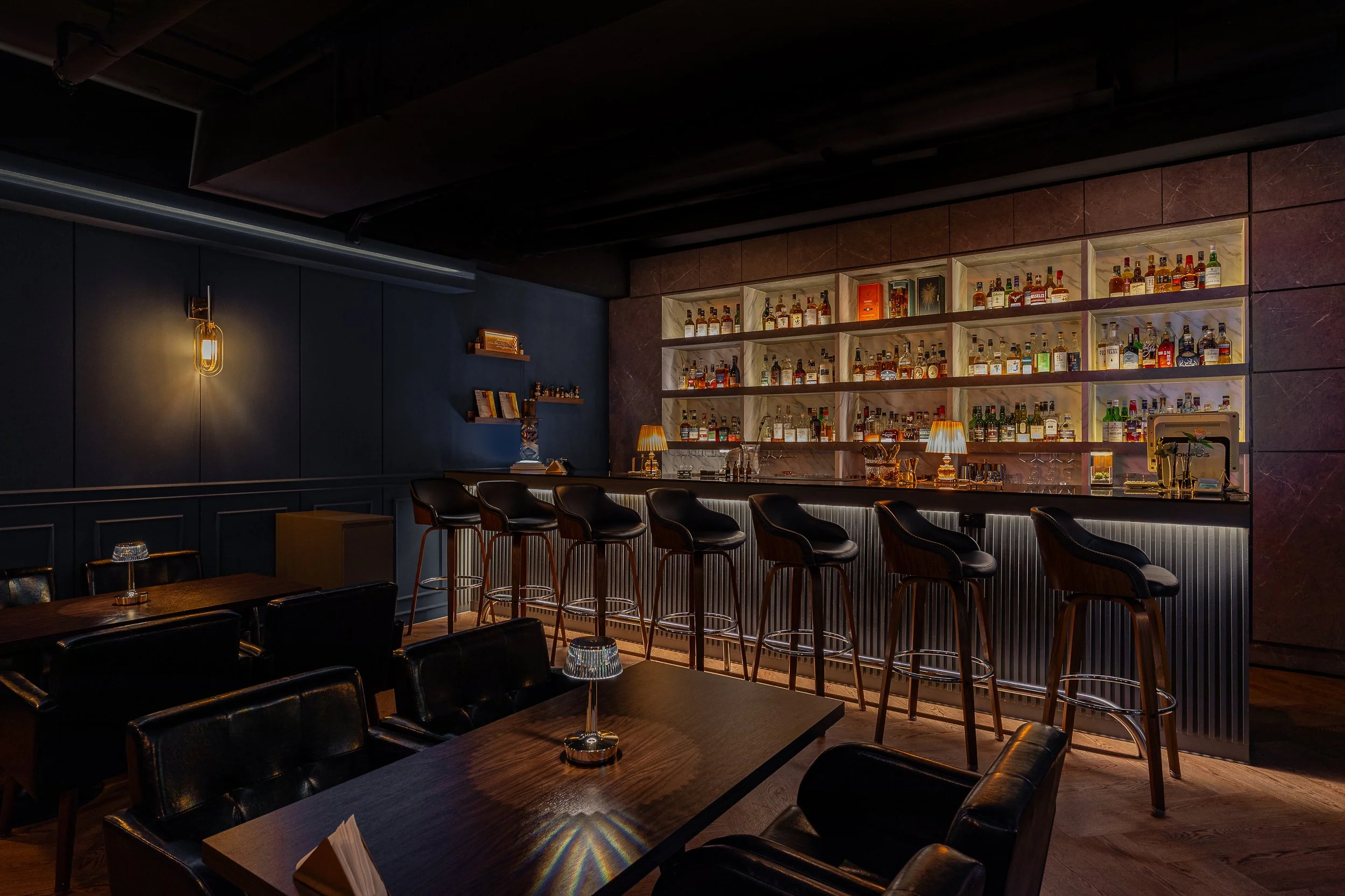 Dimly lit modern bar with black chairs and tables, illuminated shelves of liquor bottles, and warm lighting accents.