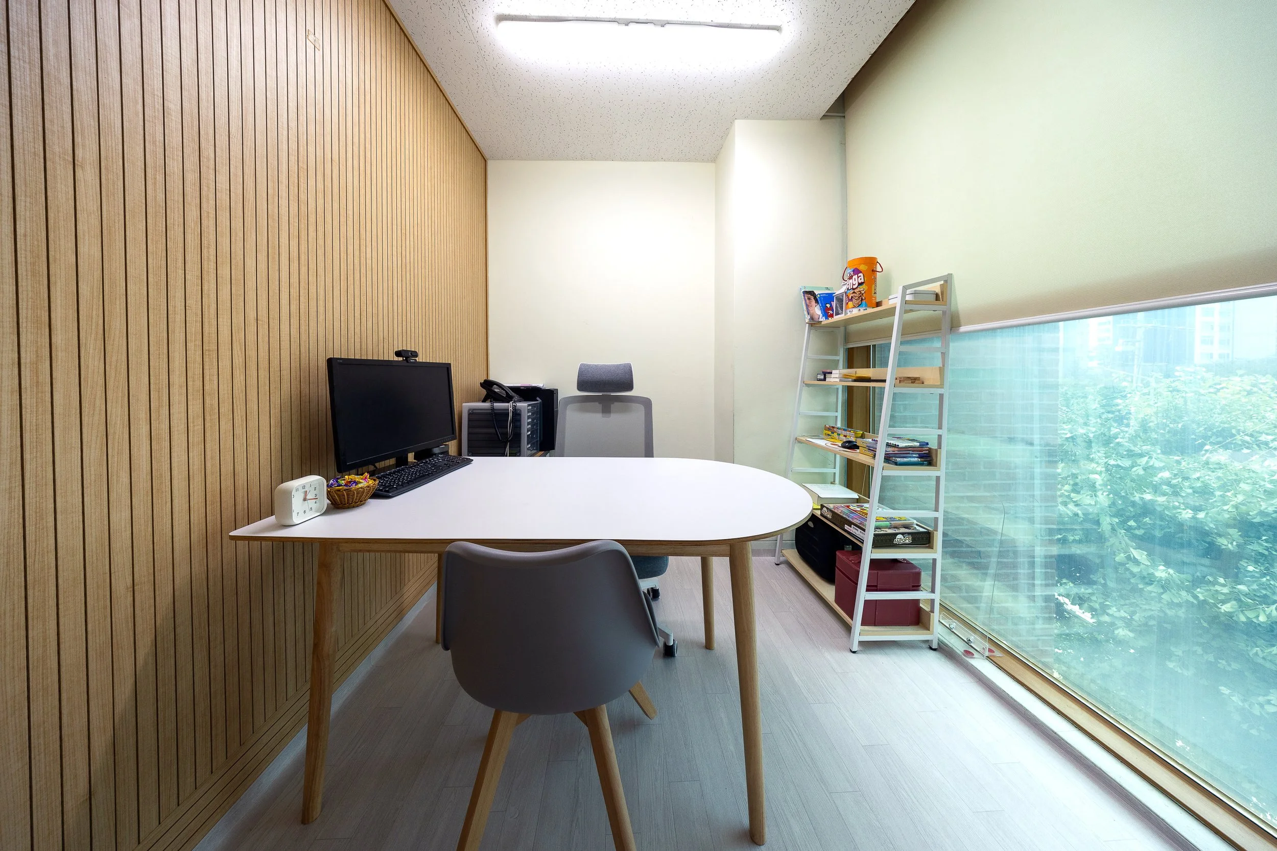 An office with a white oval desk, a computer monitor, a gray office chair, and a wooden wall. There is a bookshelf filled with books and snacks, and a large window with a view of green trees and buildings outside.