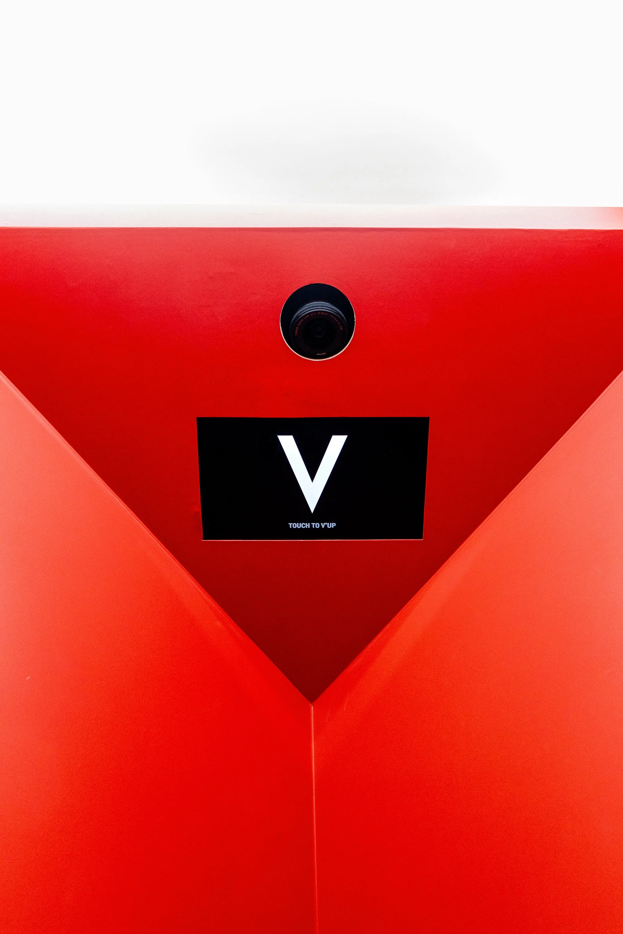 Red optical kiosk with a camera at the top and a digital screen displaying a large 'V' in white with small text below that reads 'Touch to V'up.'