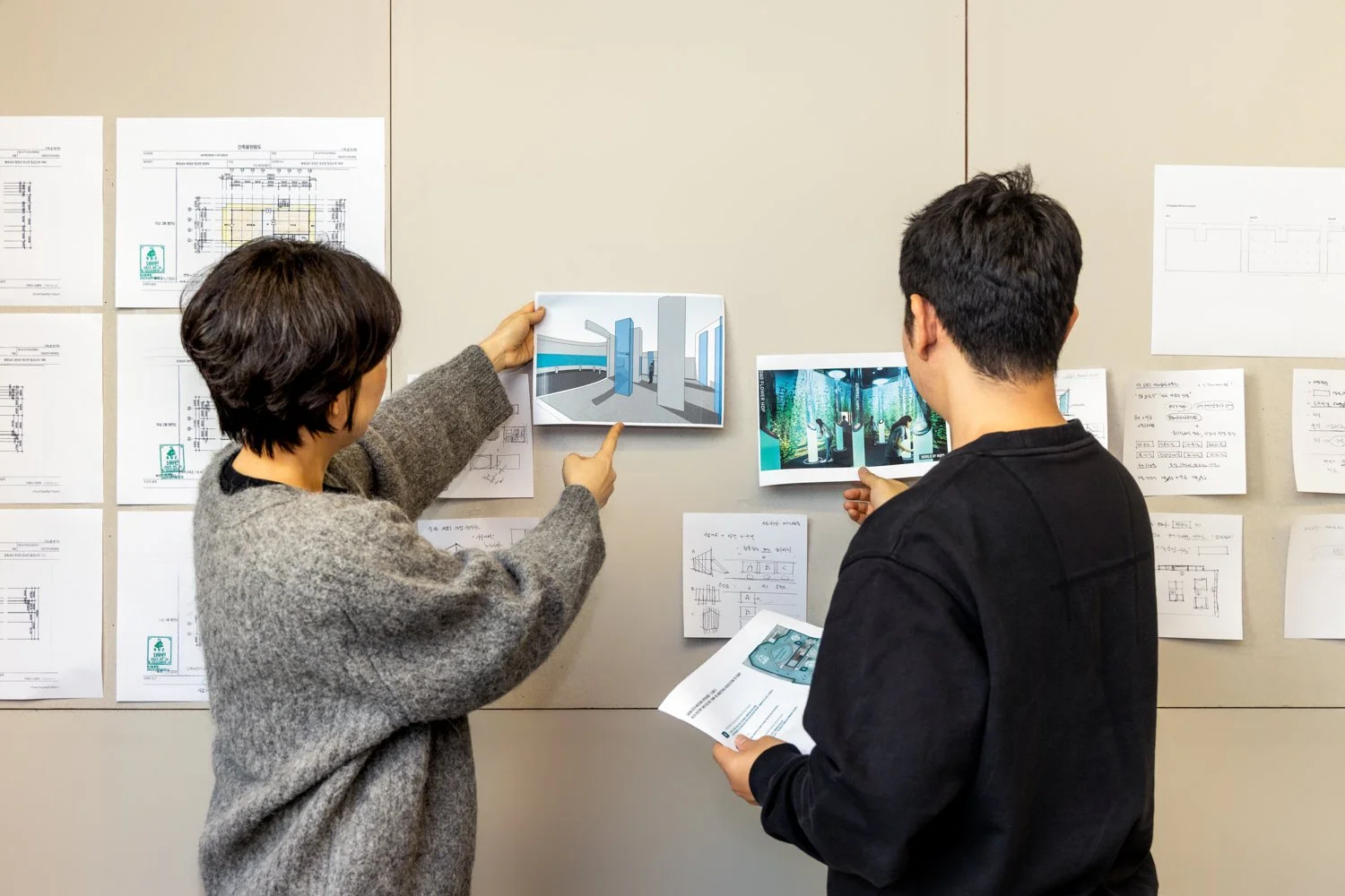 Two people discussing architectural designs in front of a wall with blueprints and printed images.