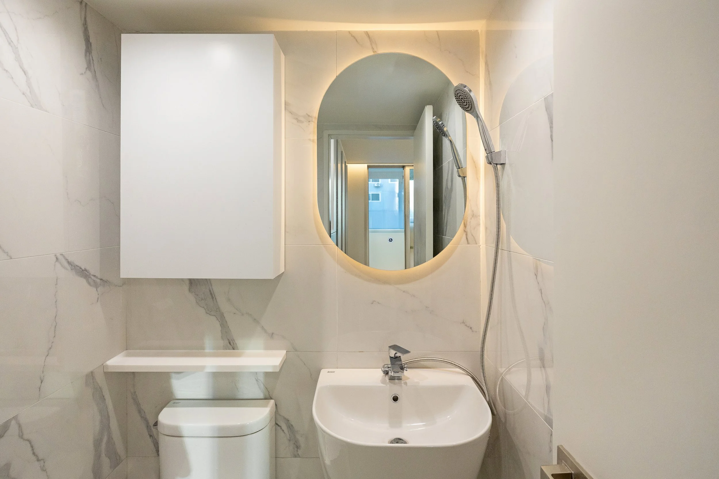 Small modern bathroom with marble walls, an oval mirror with backlighting, a white wall-mounted cabinet, a white sink, a toilet, and a handheld shower head.