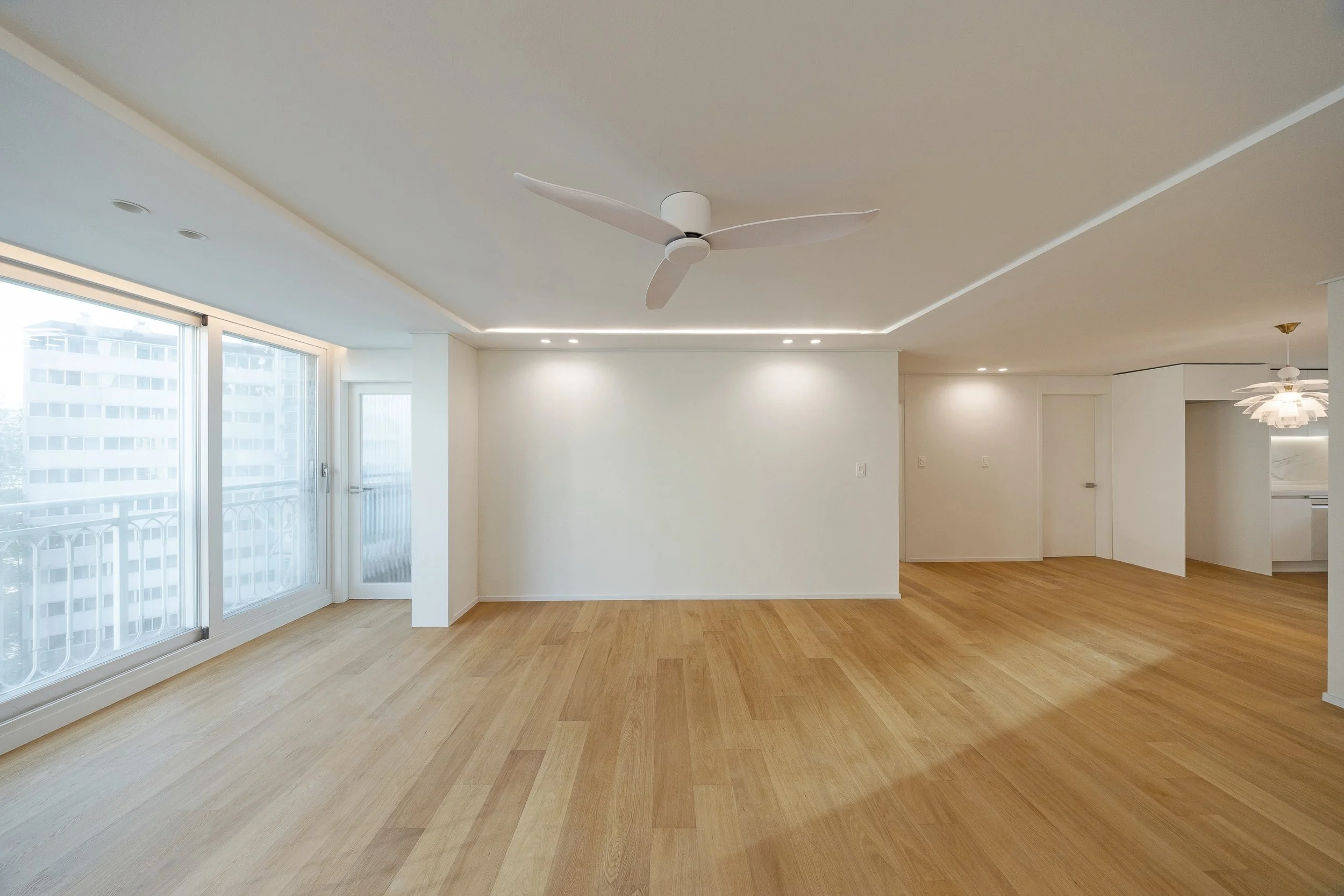 Empty living room with large windows, wooden flooring, a white ceiling fan, and modern ceiling lighting, with a view of neighboring buildings.
