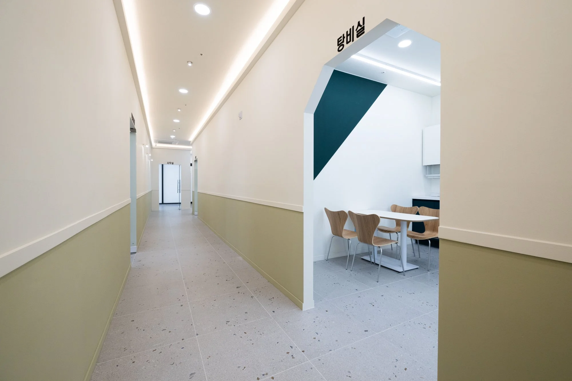 Minimalist corridor with beige walls, ceiling lighting, and a small dining area with wooden chairs and a table to the right.