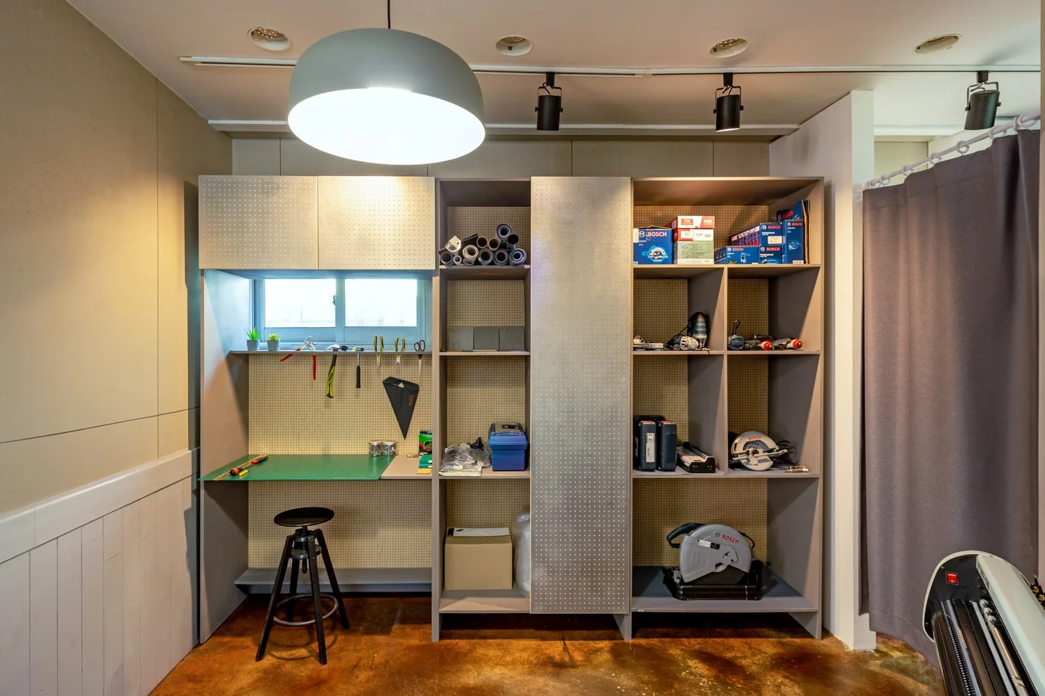 A workshop with pegboard walls, a small window, a green work surface, and shelves containing tools, boxes, and equipment. There is a stool in front of the work surface and various items hanging on the pegboard. The floor is made of reddish-brown mate