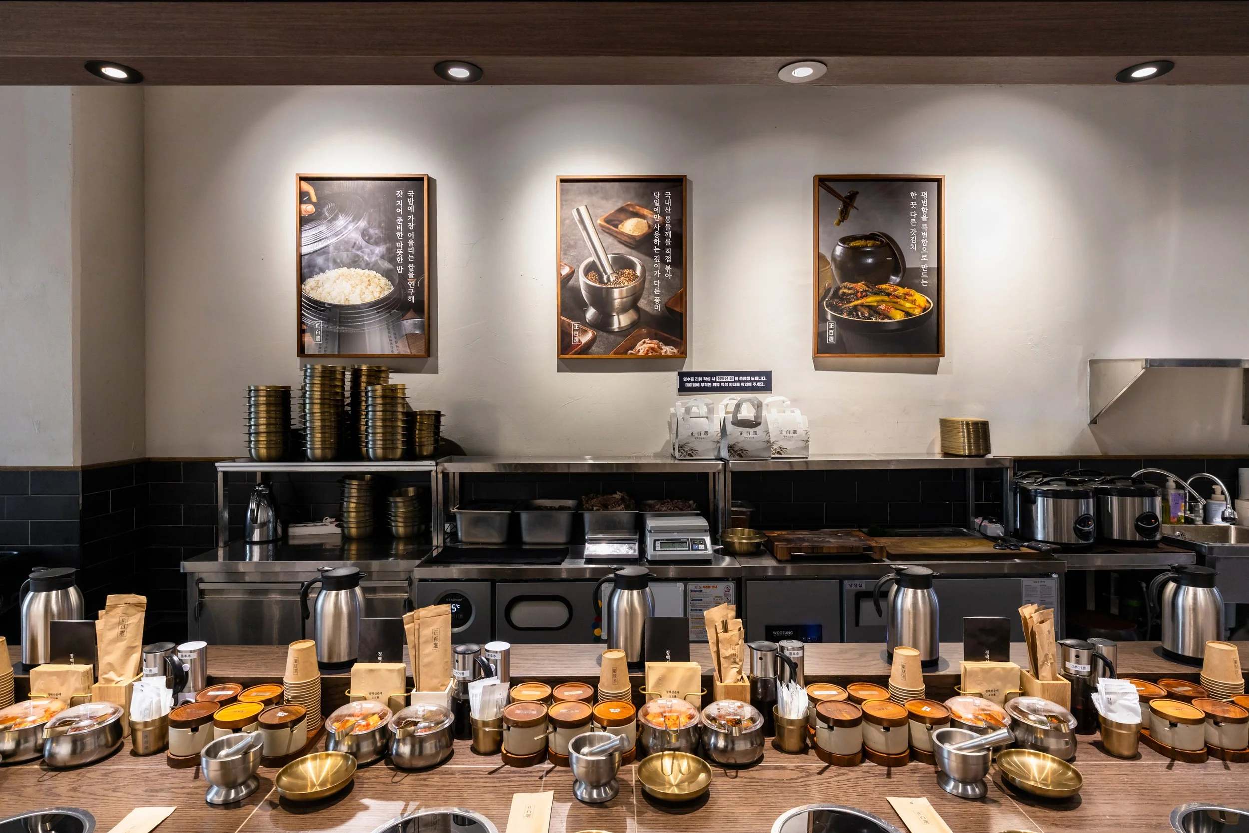 Interior of a restaurant kitchen with a counter displaying various small bowls and containers, behind which are stacked bowls, kitchen appliances, and framed food photographs on the wall.