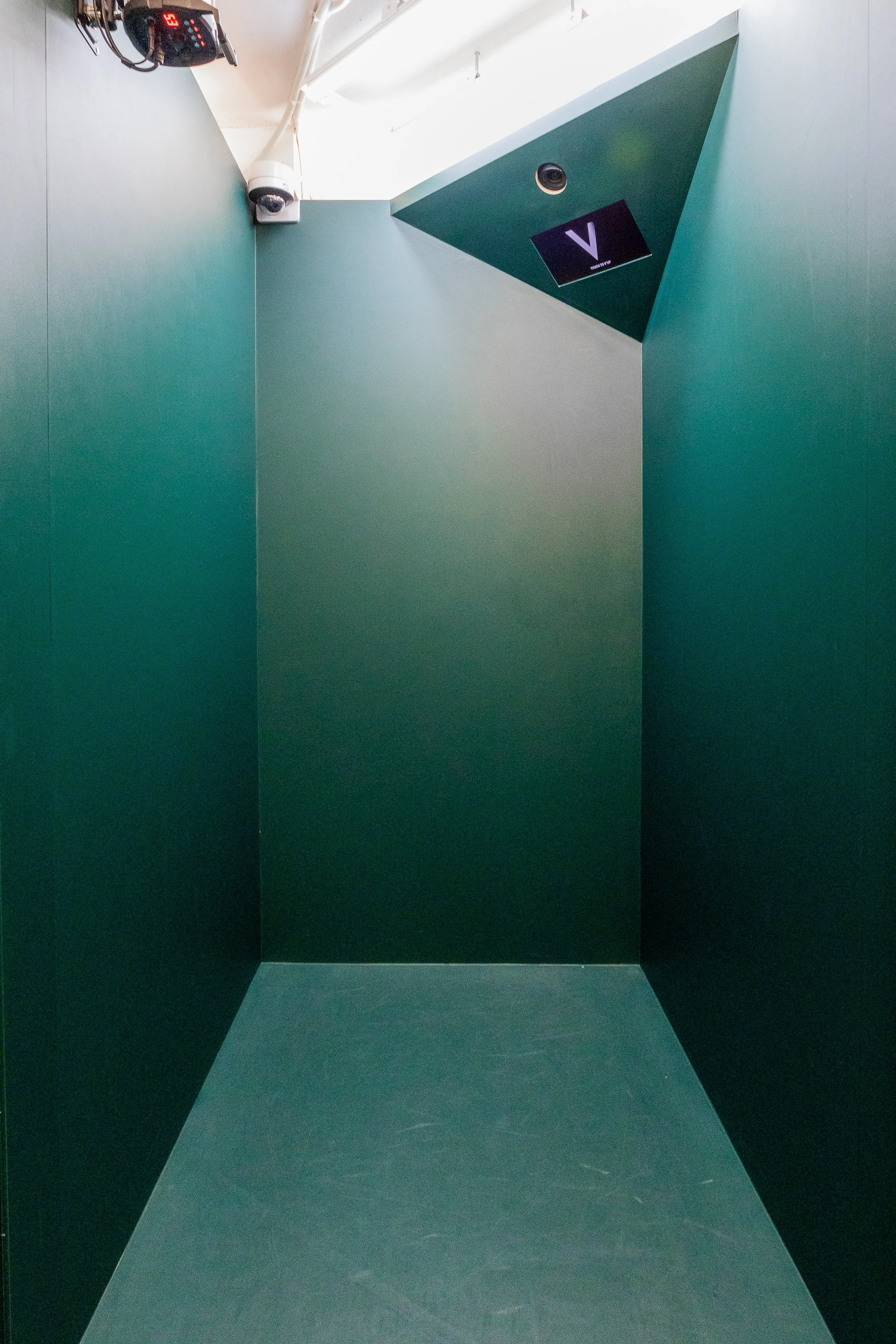 Empty green-walled elevator with a digital display showing the number '3' and a small screen with a 'V' logo on the ceiling.