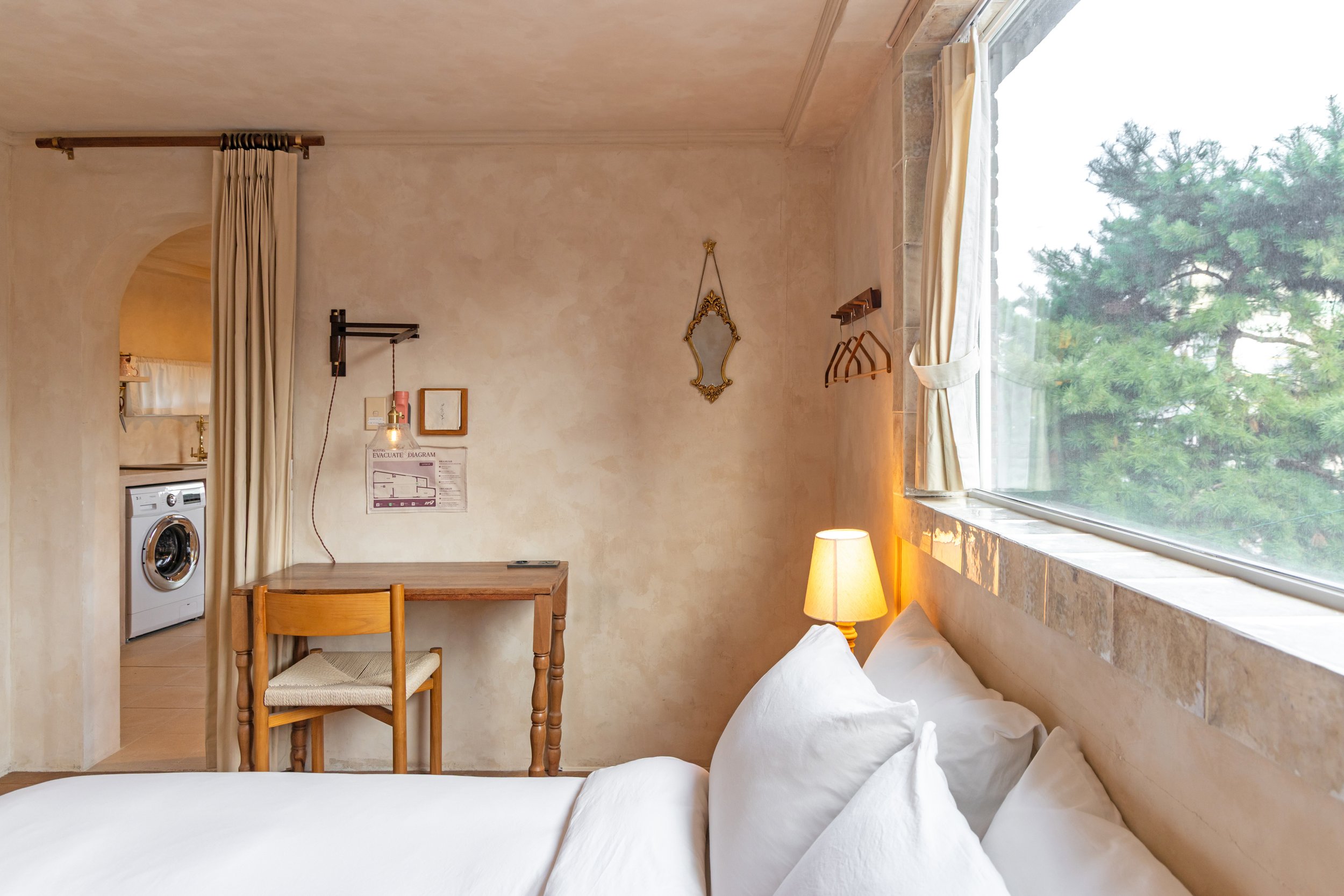 Interior view of a cozy bedroom with a bed, a window with curtains, a small wooden desk with a chair, a lamp on a nightstand, and a window showing green trees outside.