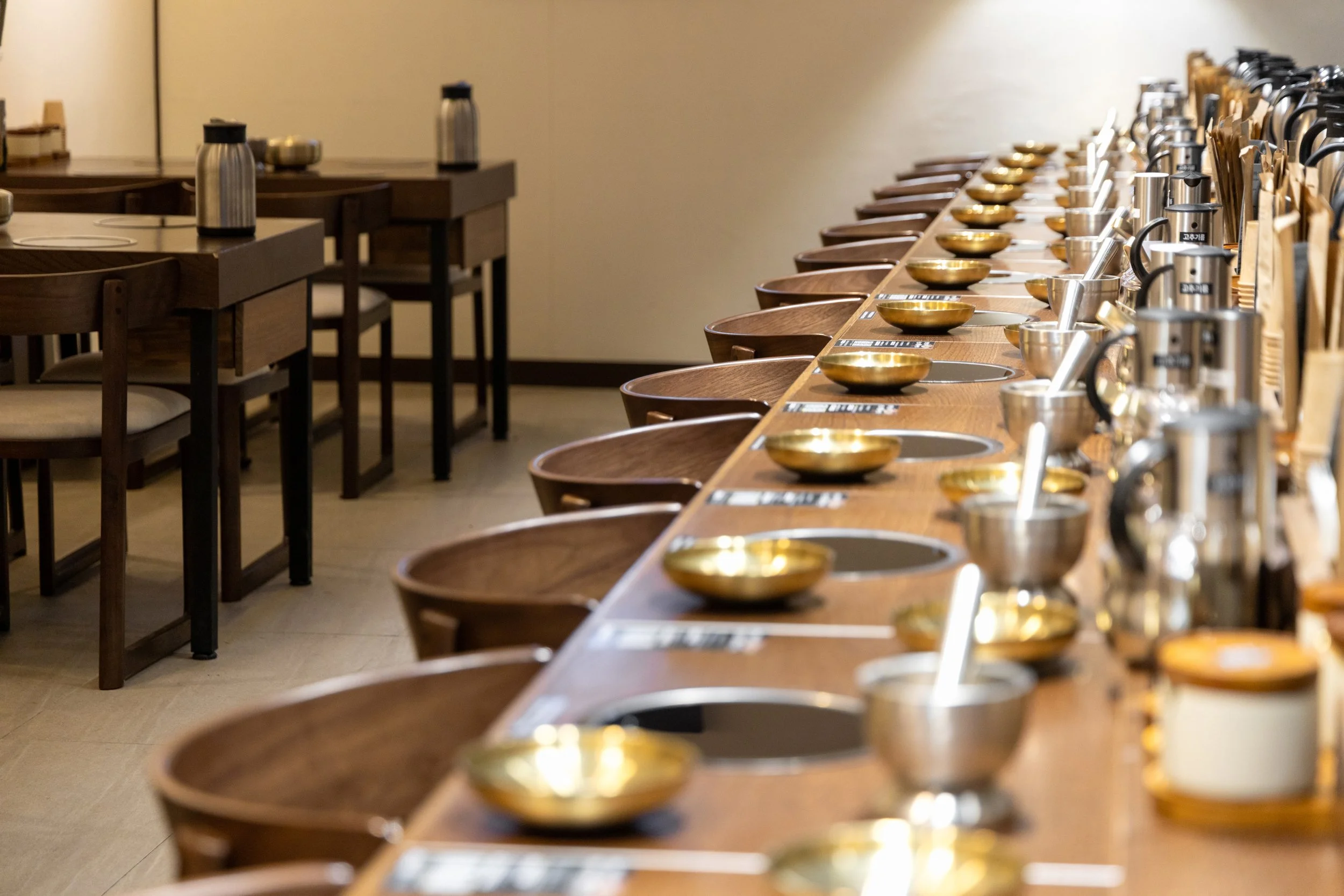 A row of wooden hot pot stations with metal bowls and ladles in a restaurant setting, with additional tables and chairs in the background.