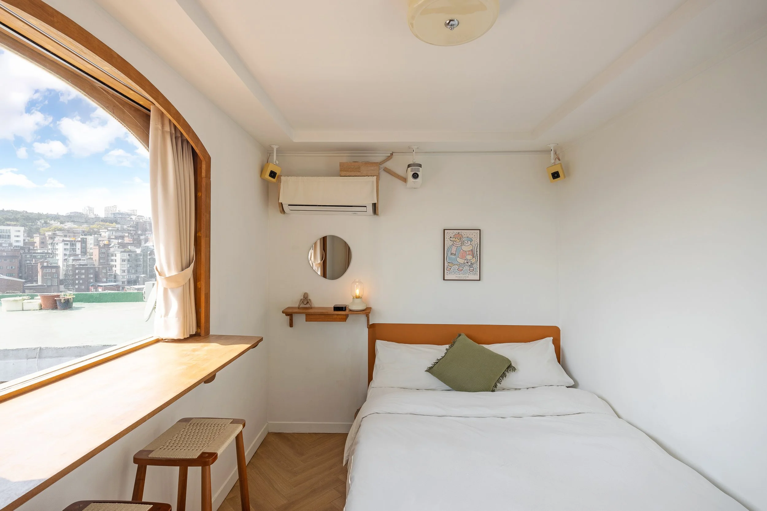 A cozy bedroom with a bed, green pillow, wall art, small floating shelf, round mirror, desk lamp, window with beige curtains, and city view outside.
