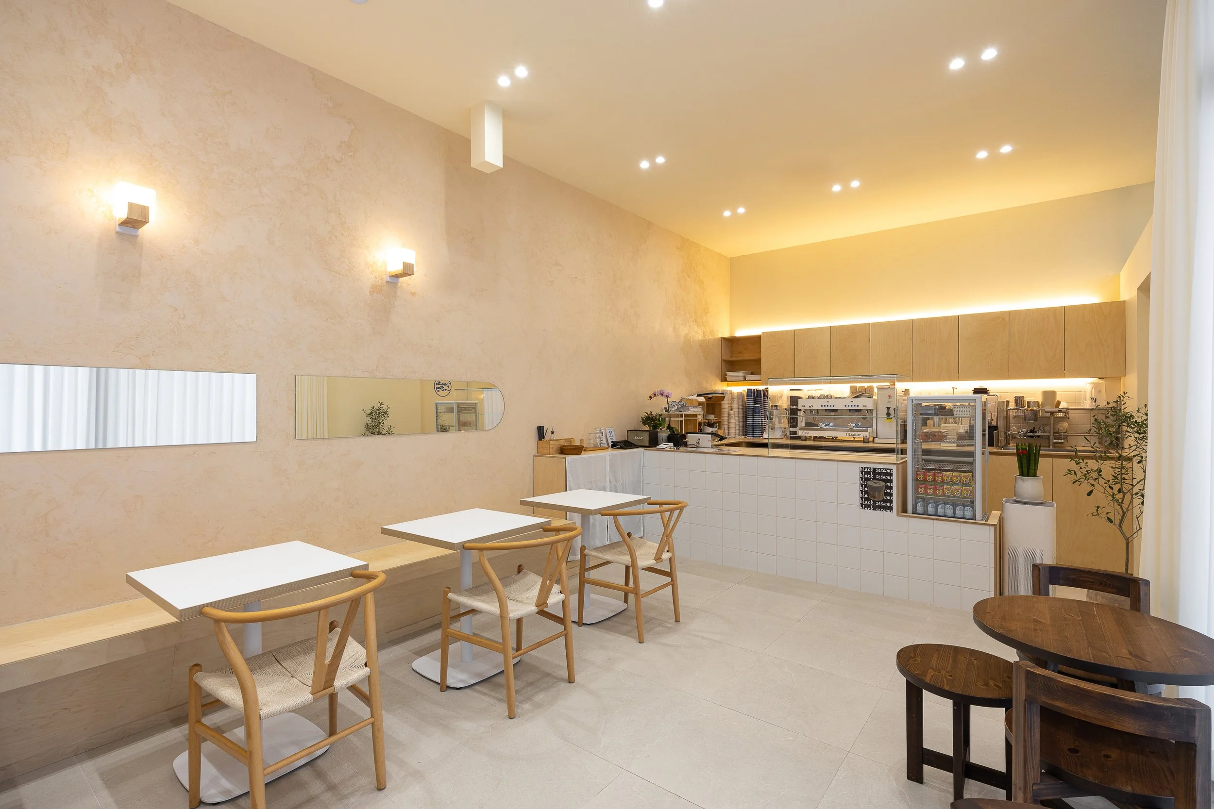 Minimalist cafe interior with white walls, beige textured wall, and warm lighting. Light wood chairs and tables near the wall, with a darker wooden table and chairs in the foreground. Coffee counter with coffee machines, cups, and a refrigerated disp