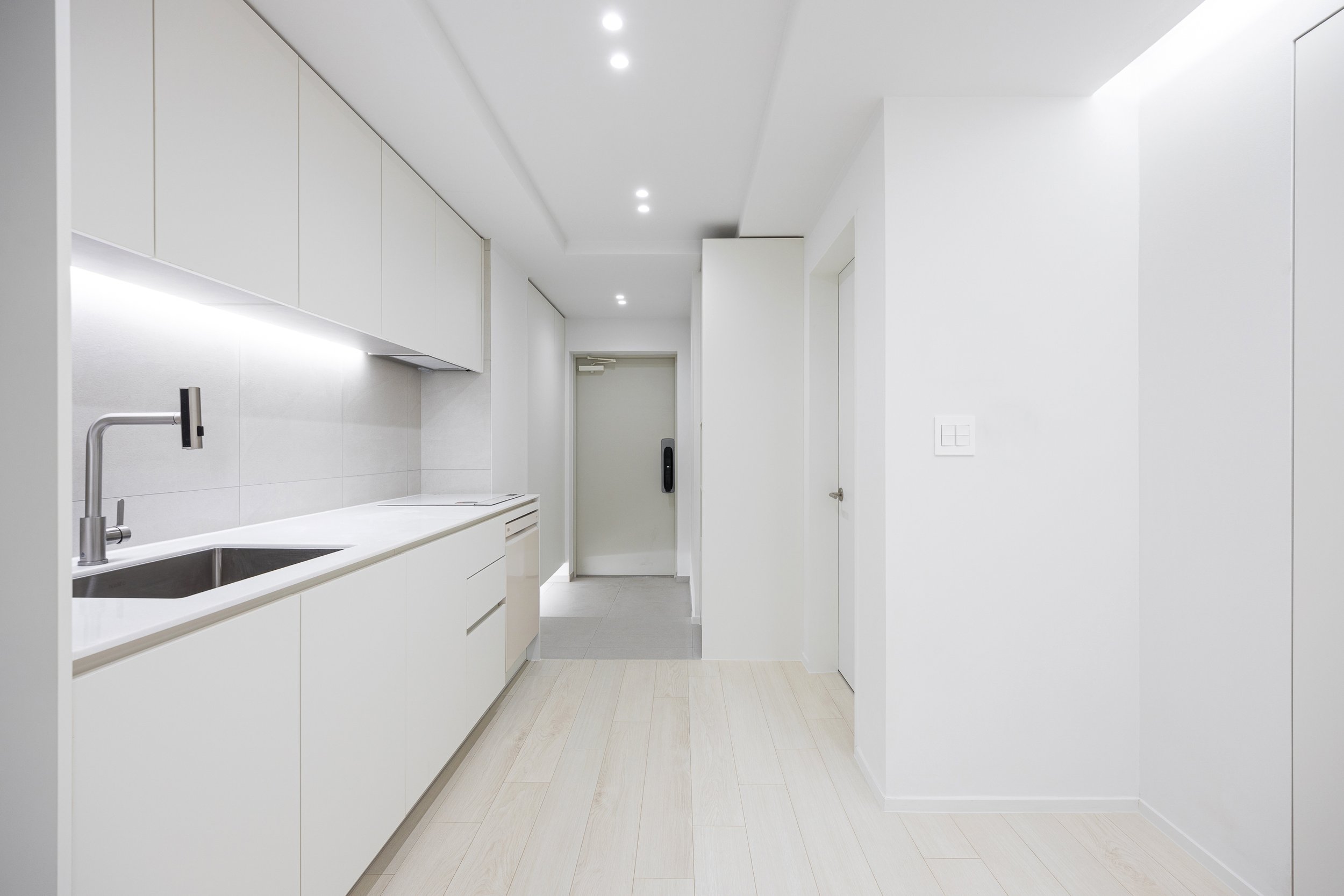 Minimalist all-white kitchen with sleek cabinetry, light wood flooring, and a view of the front door.