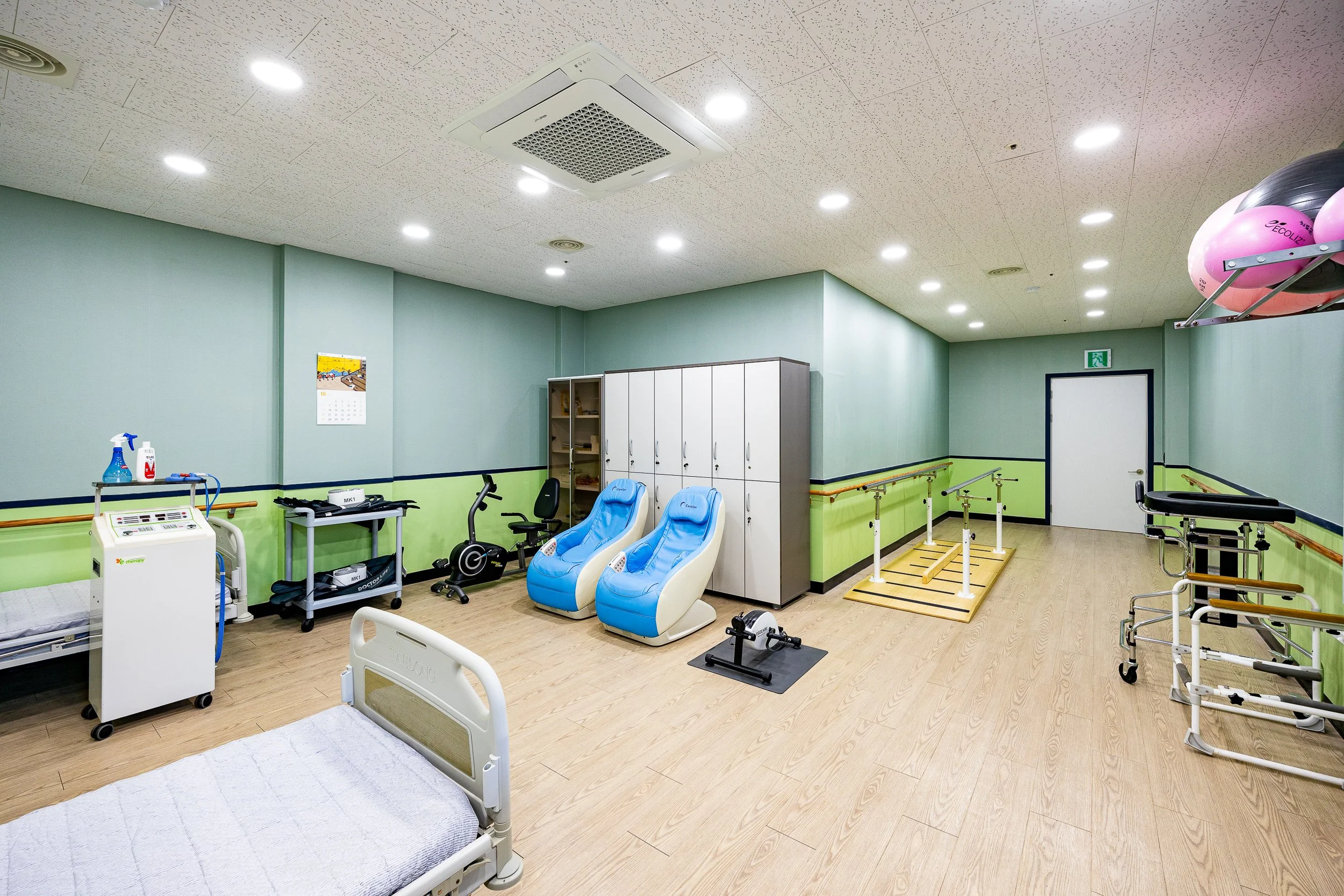 Rehabilitation therapy room with beds, exercise equipment, lockers, and therapy tools, brightly lit with overhead lights.