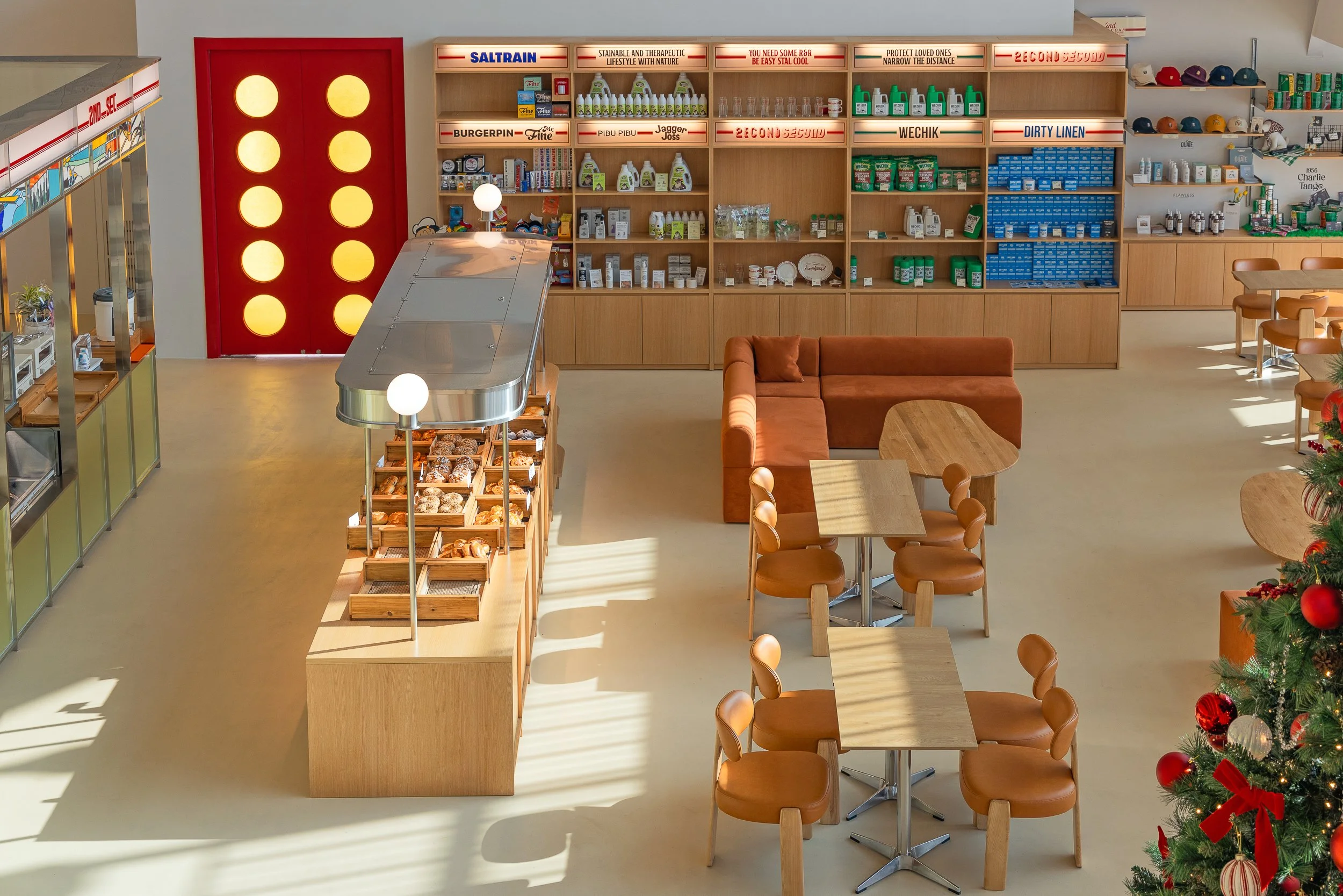 A cozy cafe or bakery interior with a display of baked goods, a seating area with wooden tables and chairs, a sofa, and a Christmas tree decorated with red ribbons and ornaments.