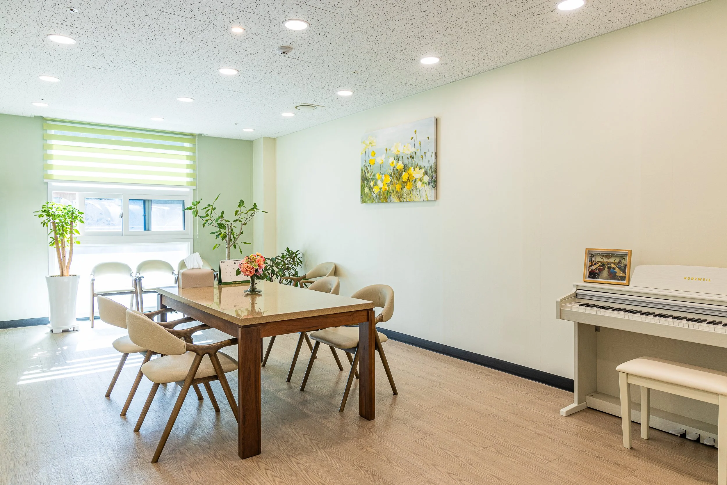 A bright room with a wooden table surrounded by beige chairs, potted plants near the window, a flower arrangement, wall art with yellow flowers, and a white upright piano with a framed picture on top.