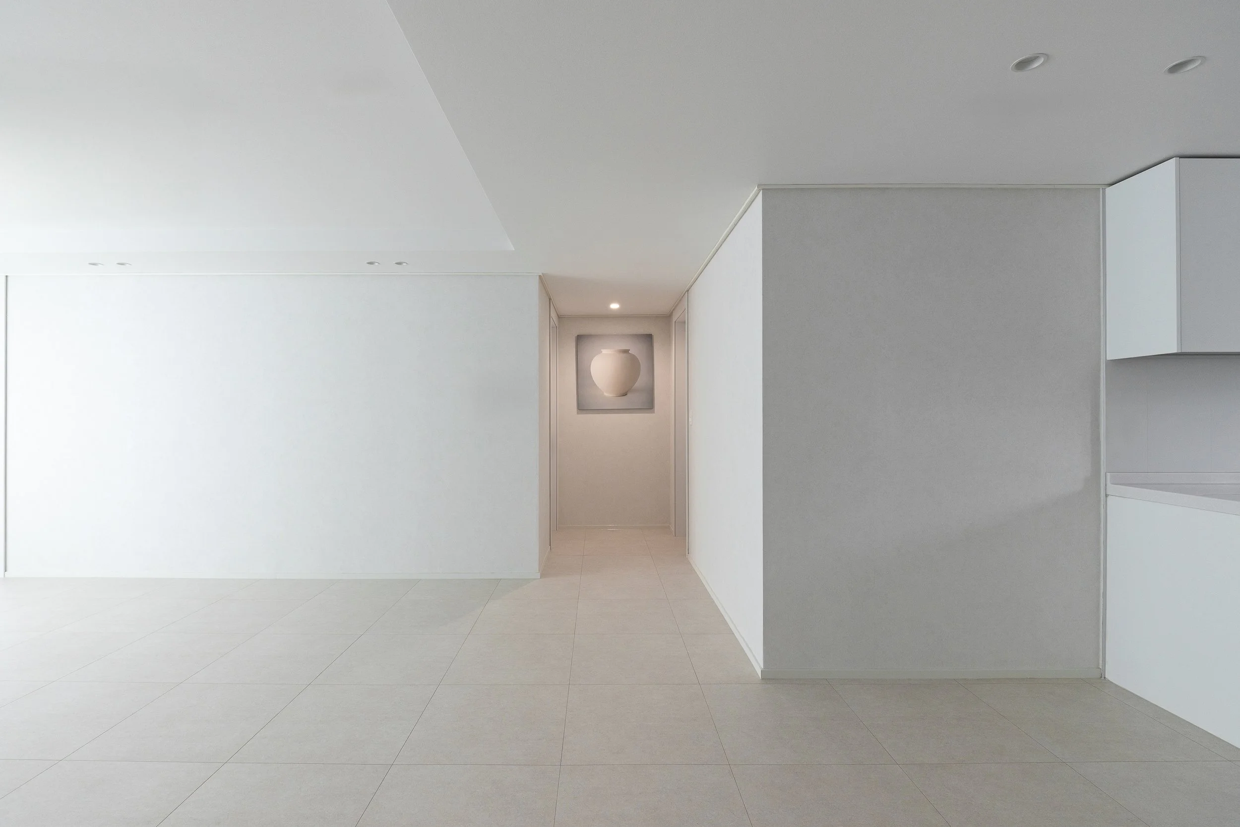 Minimalist white interior with tiled floor, plain walls, and a framed artwork of a white vase in a hallway.