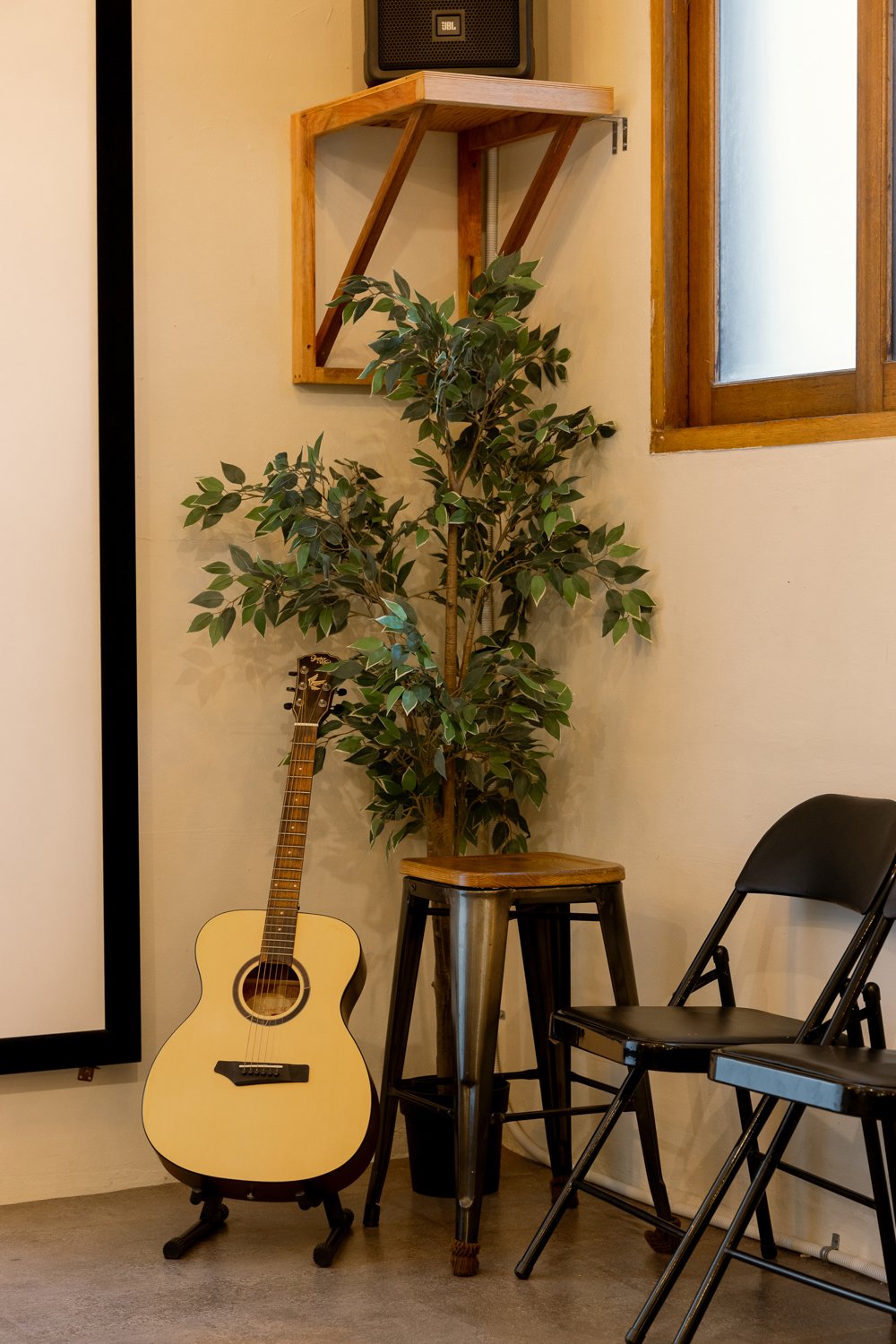 An interior scene with a wooden stool, a black folding chair, a potted leafy plant, an acoustic guitar, a small wooden shelf mounted on the wall, a window with a wooden frame, and a speaker on the shelf.