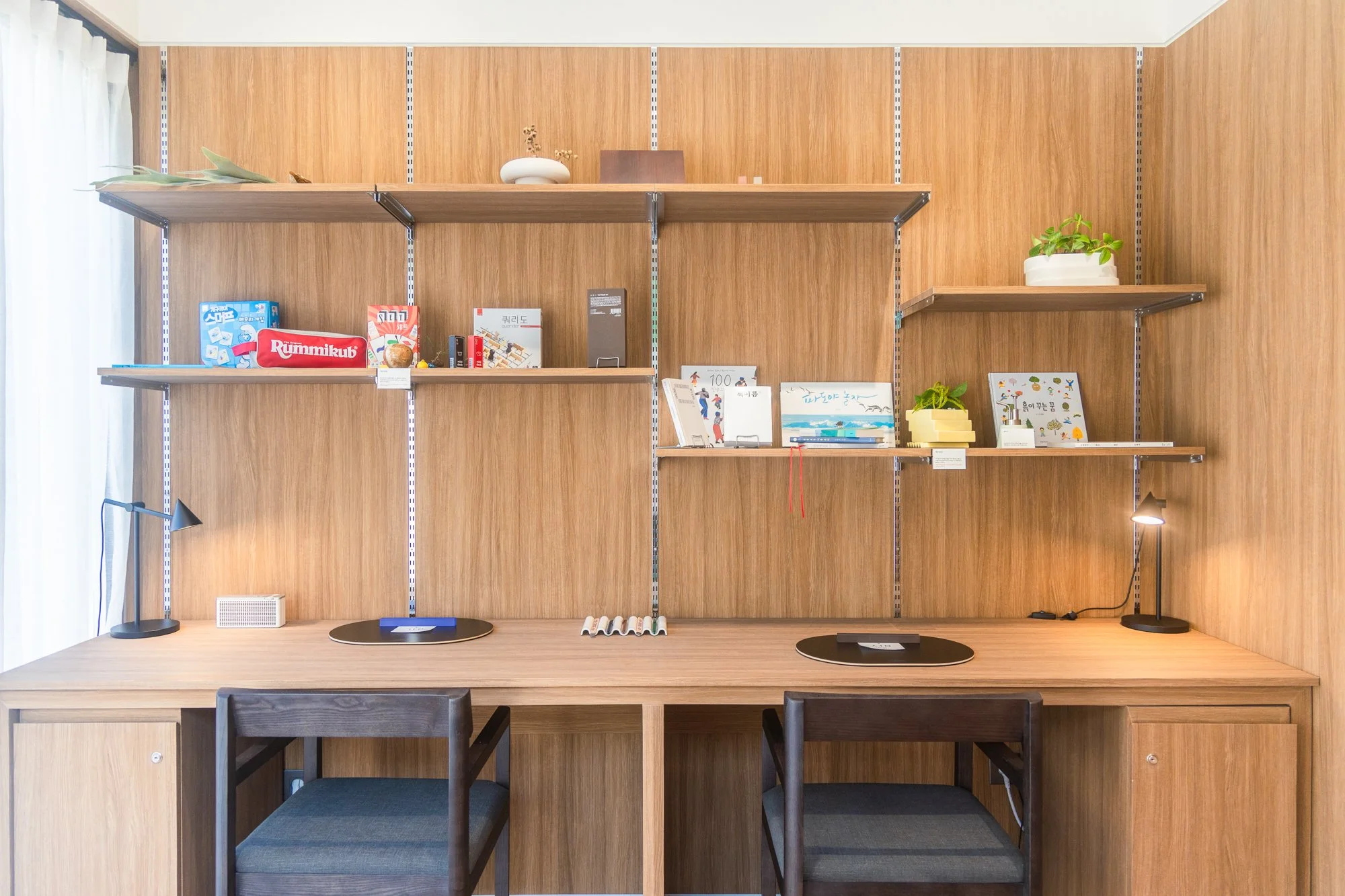 Wooden desk with two chairs, two black desk lamps, and a small speaker, set against a wooden wall with shelves holding books, plants, and decorative items.