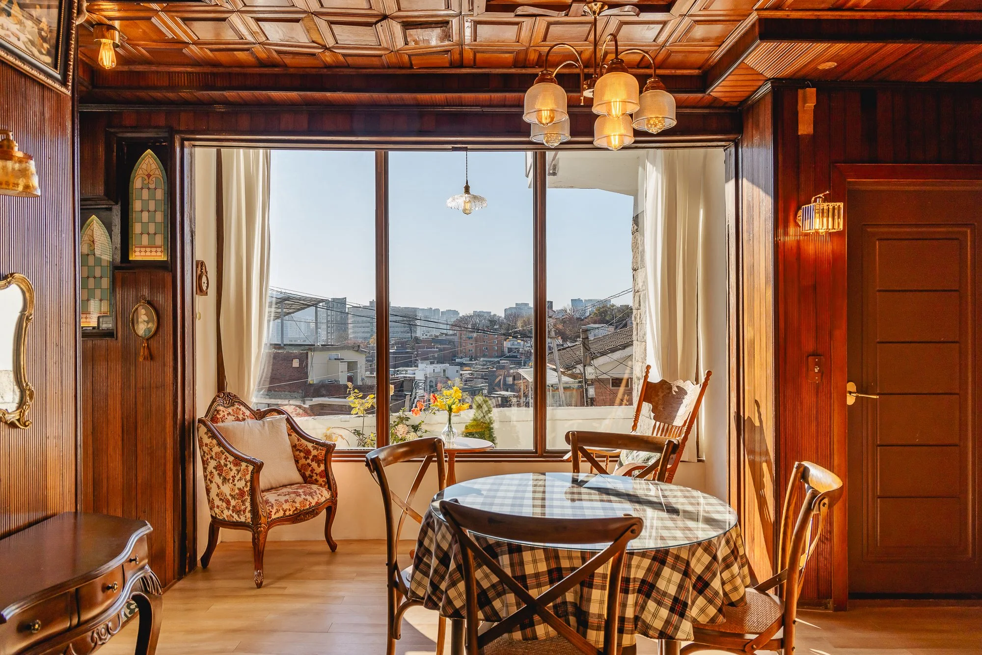 Sunlit dining area with round table covered with a checkered tablecloth, surrounded by four wooden chairs, in front of large window with view of city buildings and rooftops, wood-paneled walls, ornate upholstered armchair, and decorative pendant and 