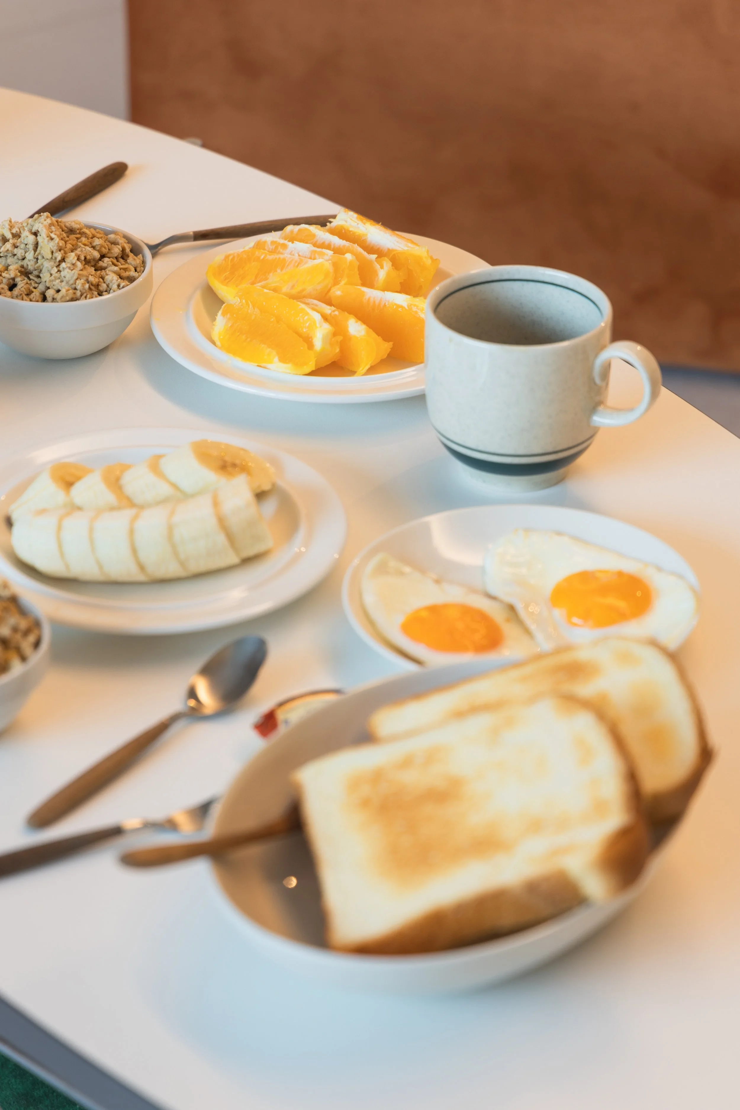 Breakfast with fried eggs, toast, bananas, orange slices, coffee, cereal, and a bowl of oatmeal or granola on a white table.
