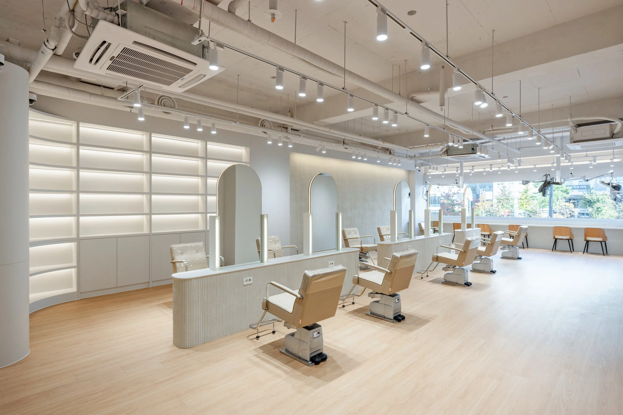 Empty beauty salon with styling chairs, mirrors, and large windows, bright lighting, modern minimalist decor, and wood flooring.