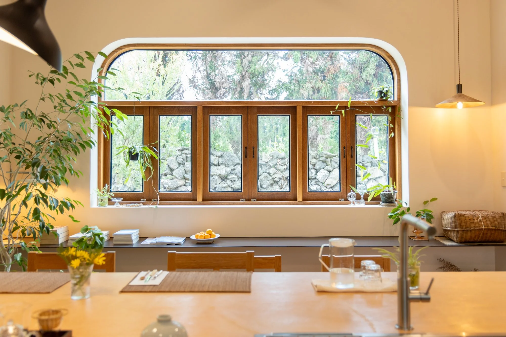 Bright kitchen with large arched window, wooden framed smaller windows, potted plants, and a wooden dining table with place settings and a glass pitcher of water.