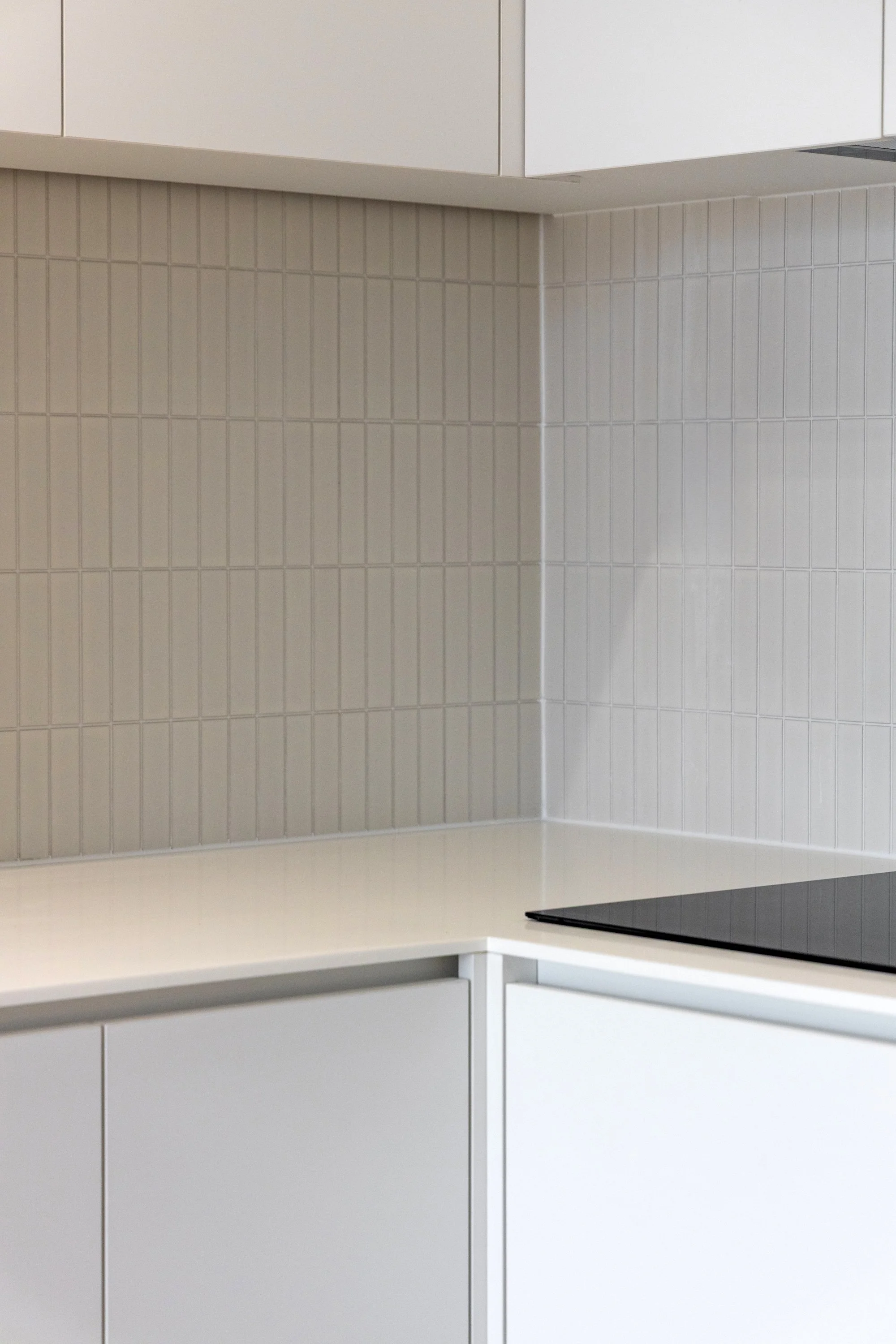 A corner of a modern white kitchen with white cabinets, beige tiled backsplash, and a black electric cooktop.