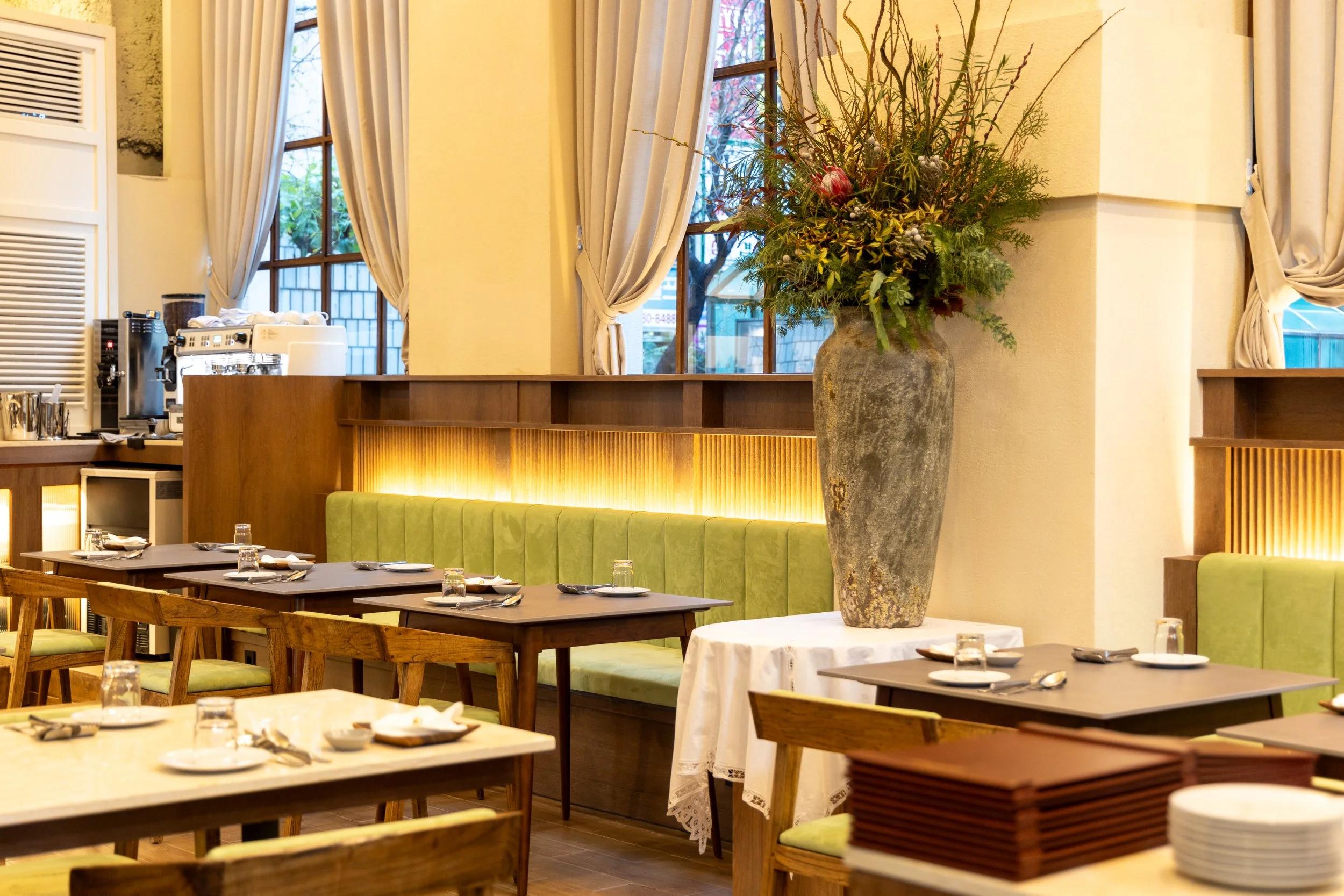 Indoor dining area with tables set with plates, glasses, and cutlery. Large flower arrangement in a tall vase on a small table. Green cushioned seating along the wall, with windows and cream curtains in the background.