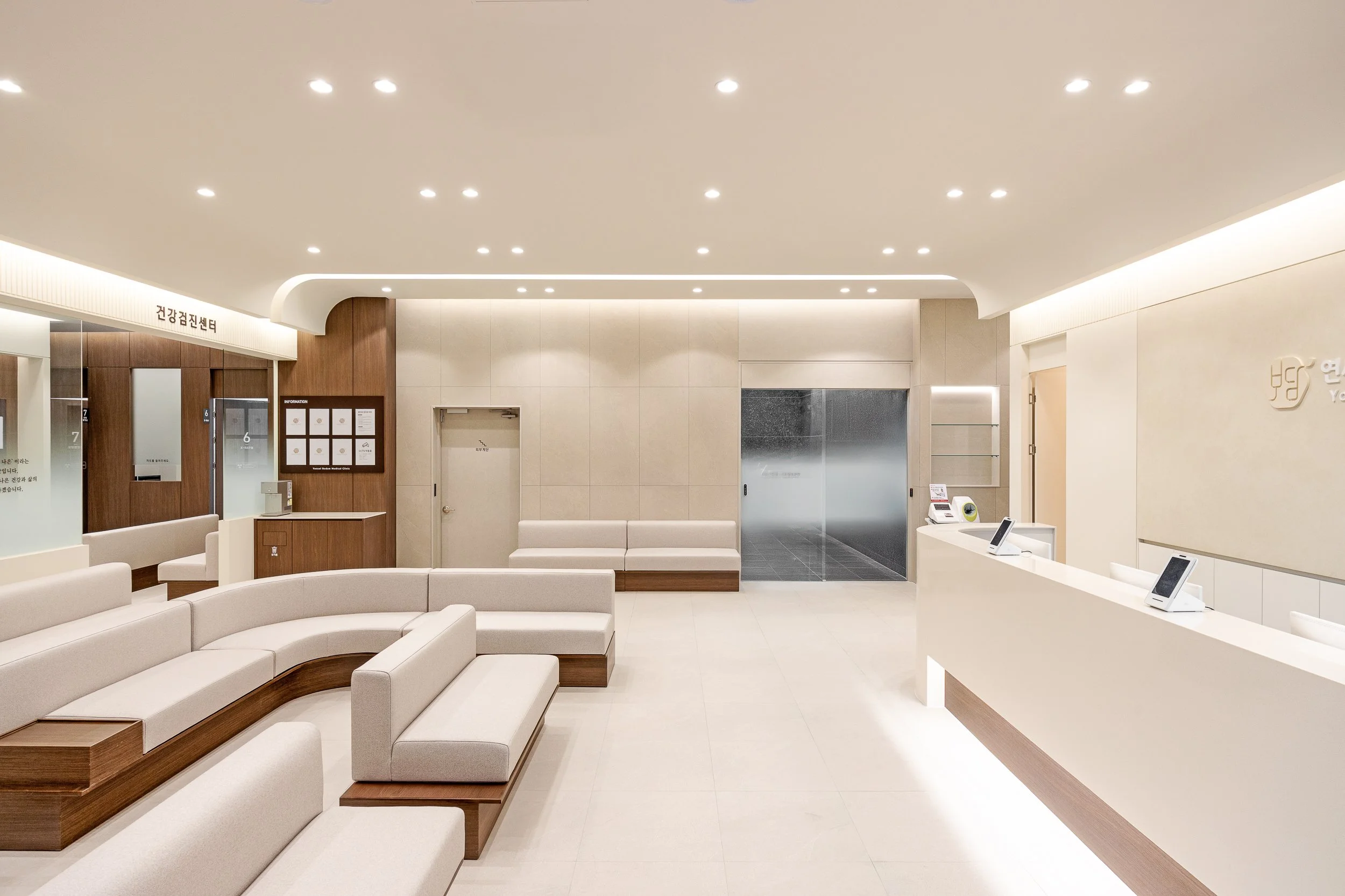 Modern, minimalist hospital or clinic waiting room with white seating, wooden accents, and a reception desk with digital tablets.