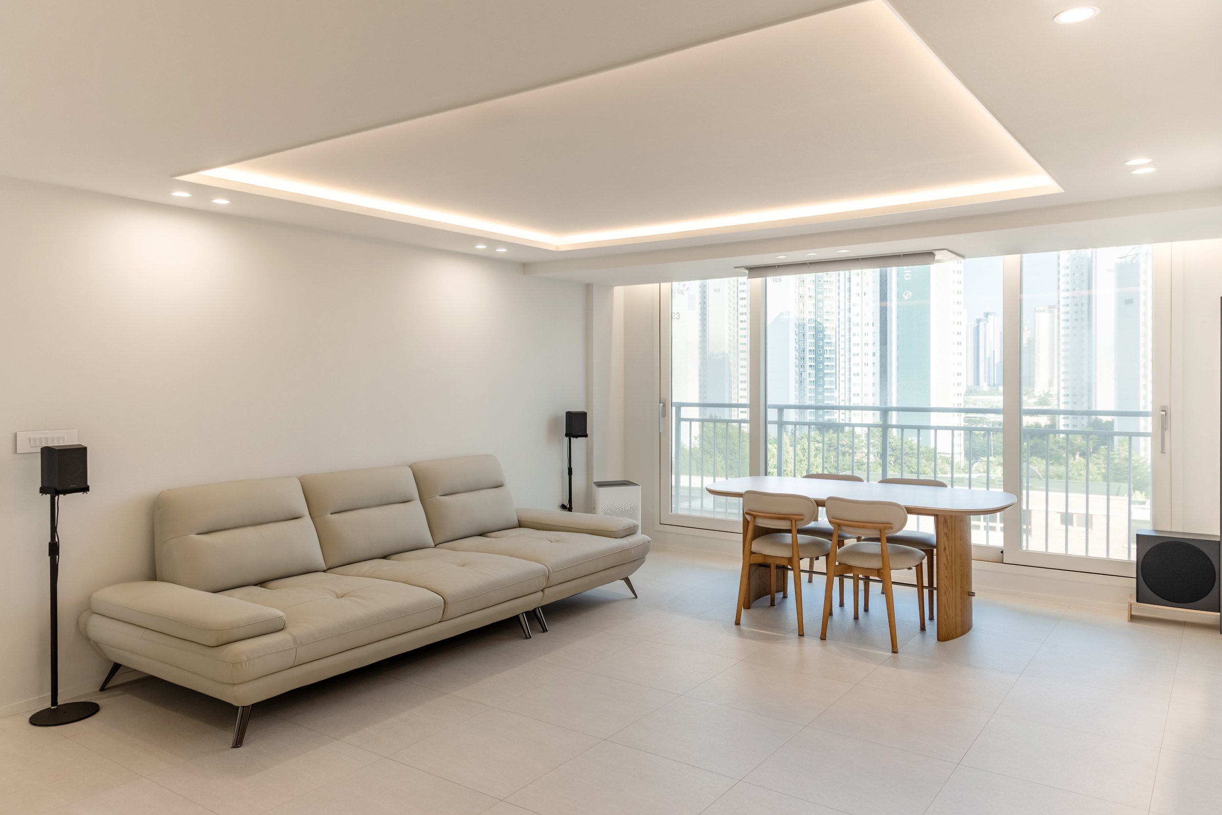 Modern living room with a cream sofa, two black speakers, a wooden dining table with three chairs, large sliding glass doors leading to a balcony, and city skyline views.