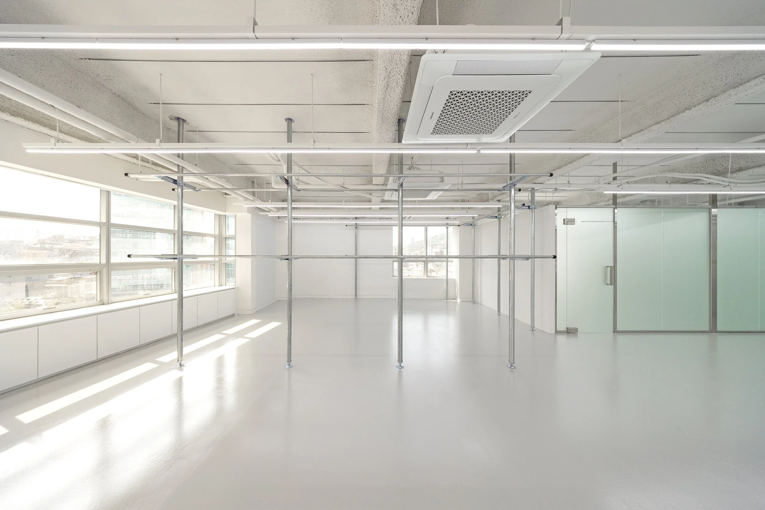 Empty modern office space with large windows, white floors, and metal scaffolding in the center.