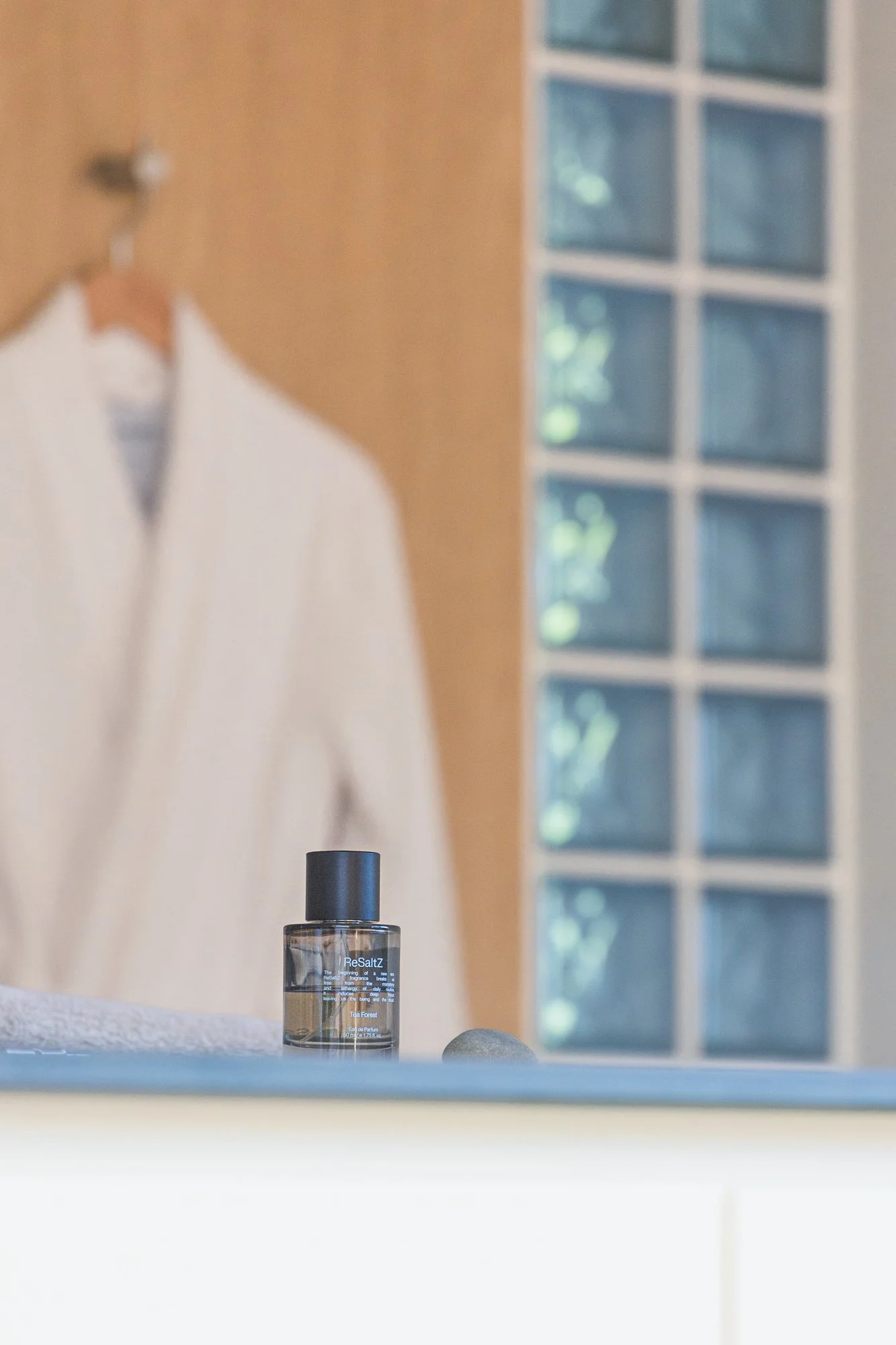 A small bottle of ReSaltZ on a bathroom counter with a white towel and a gray stone nearby, reflected in a mirror that shows a blurred person's white shirt and a glass block window.