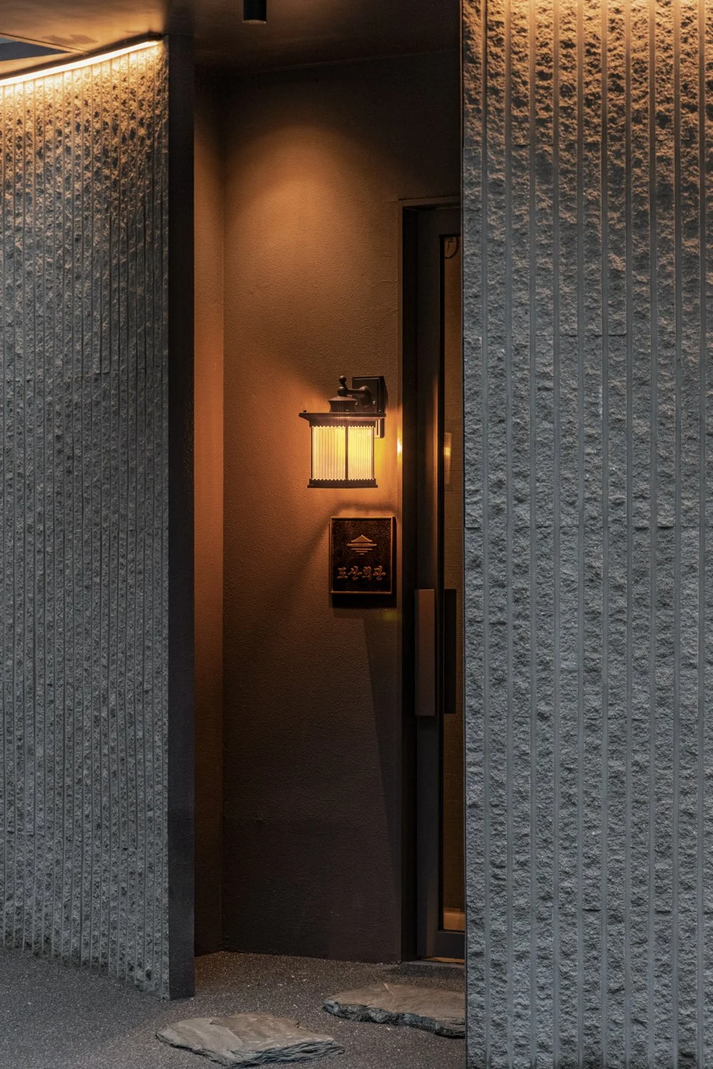 Nighttime shot of a modern exterior entryway with a glowing wall-mounted lantern, a doorbell, and a vertical window framed by textured gray concrete walls.