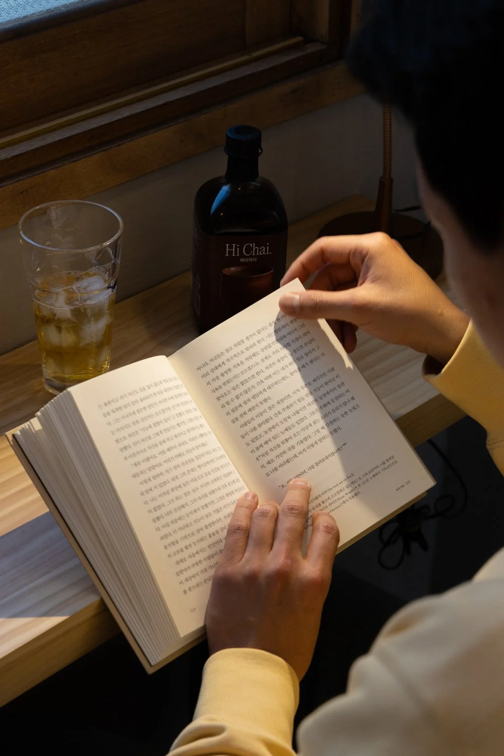 A person reading a book at a wooden table with a glass of iced tea and a bottle labeled "Hi Chai" nearby.