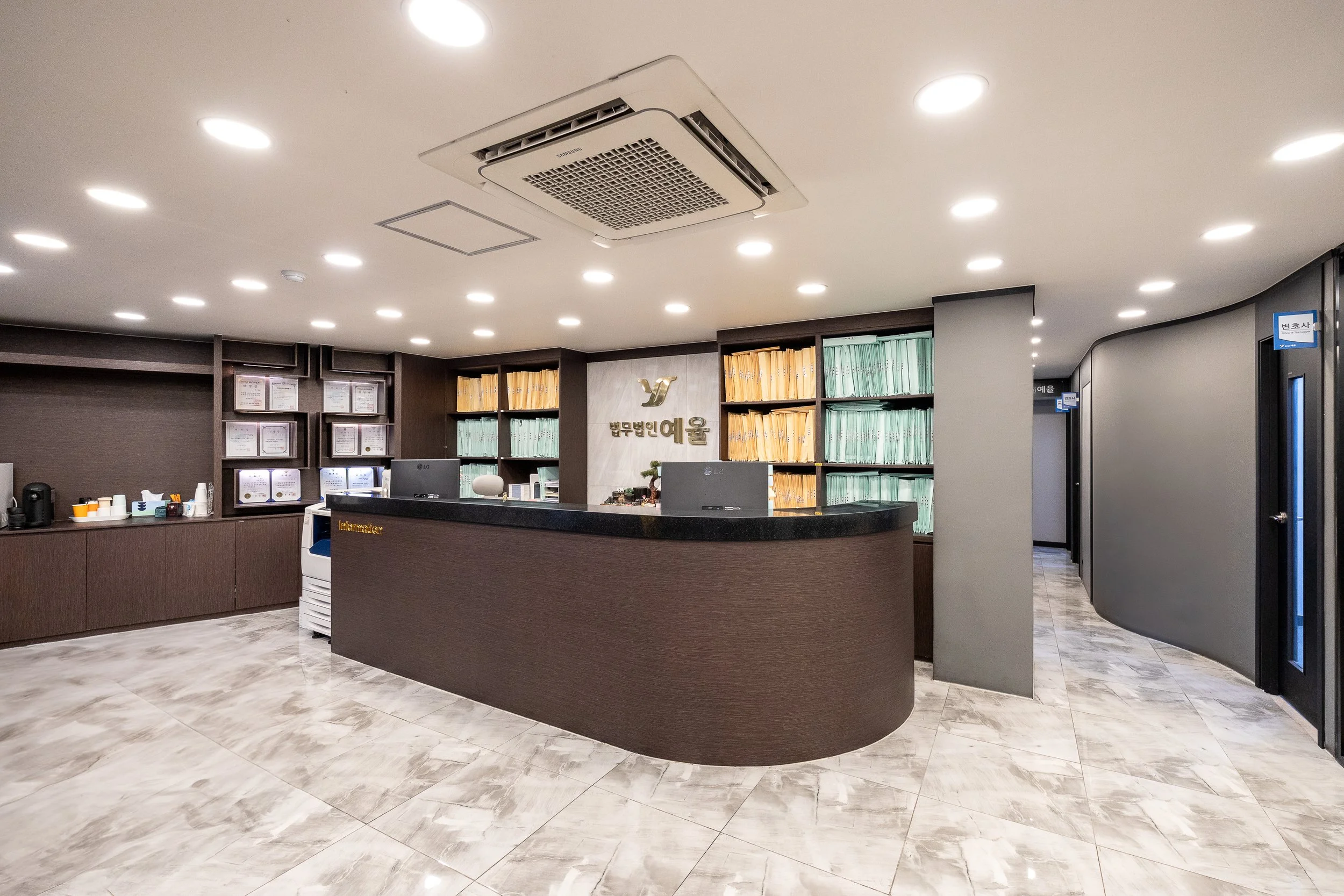 Modern reception area with a curved dark wood counter, surrounded by shelves with yellow and green files, and a wall with framed certificates and a logo in Korean.