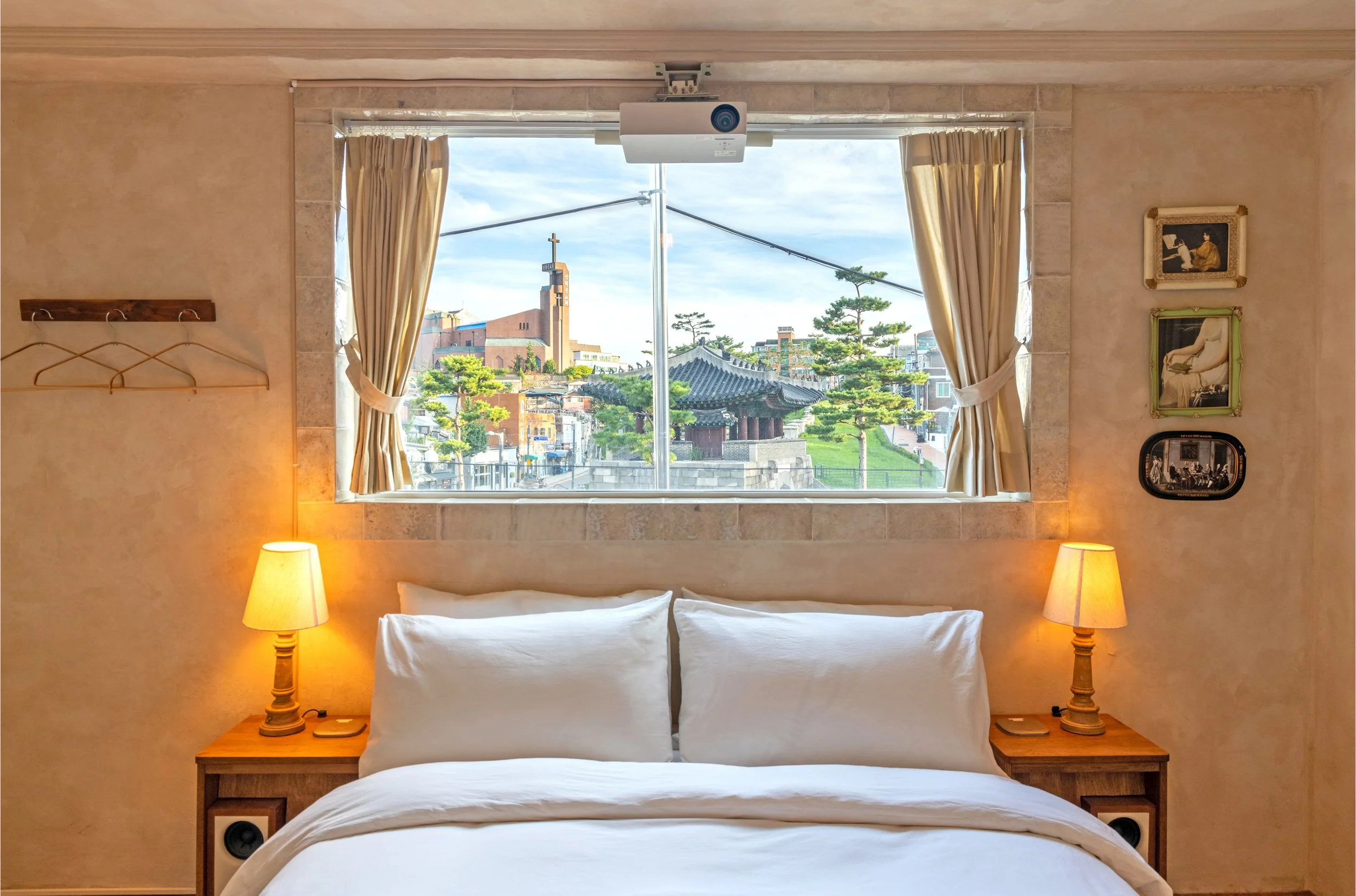 Bedroom with a large window showing a cityscape, a traditional building, and trees outside. The bed has white bedding, and there are two bedside lamps turned on. The room has beige walls with framed pictures and a wall-mounted coat rack on the left.