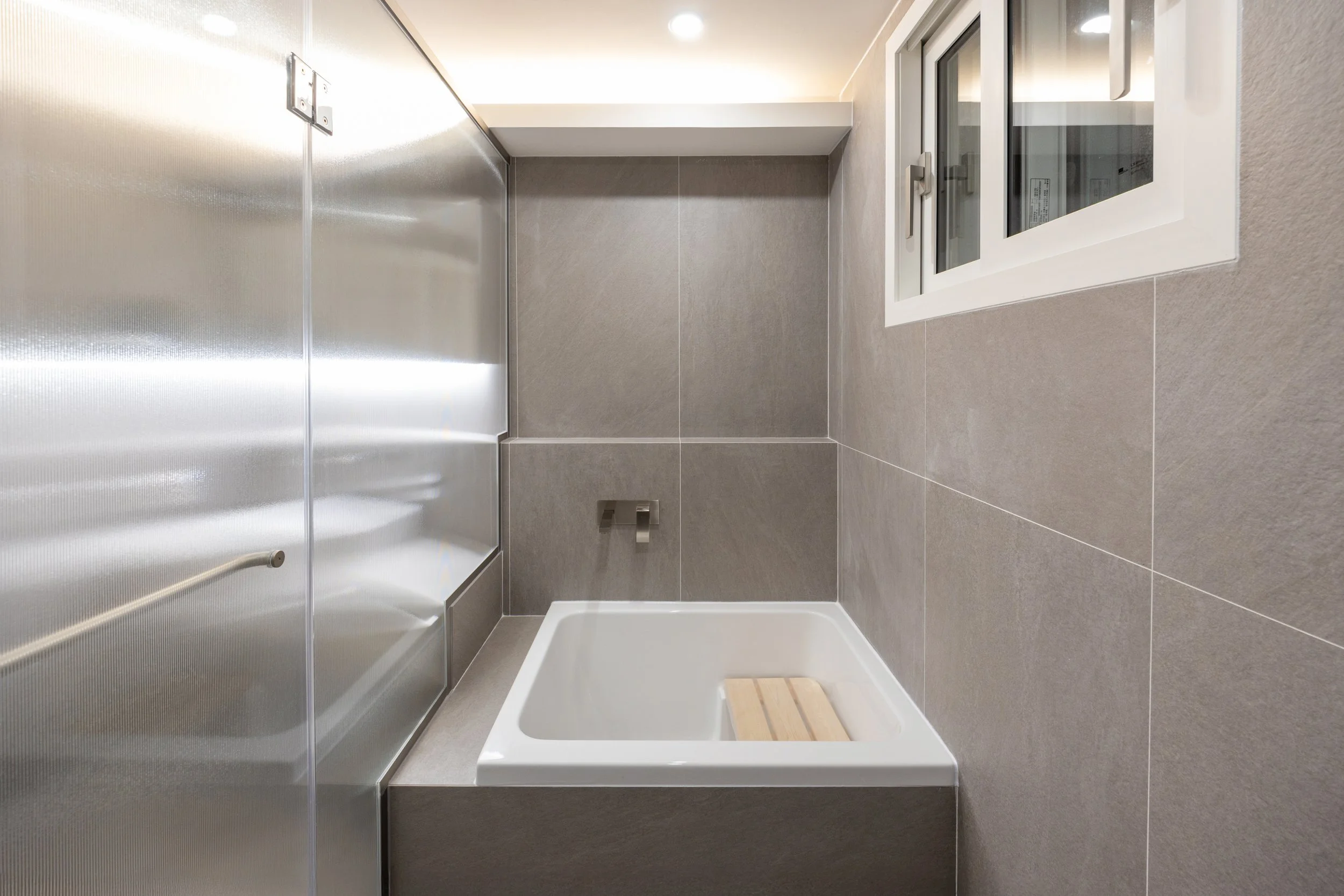 Modern bathroom with gray tiled walls and floor, white bathtub with wooden bathmat, stainless steel shower enclosure, and a large sliding window.