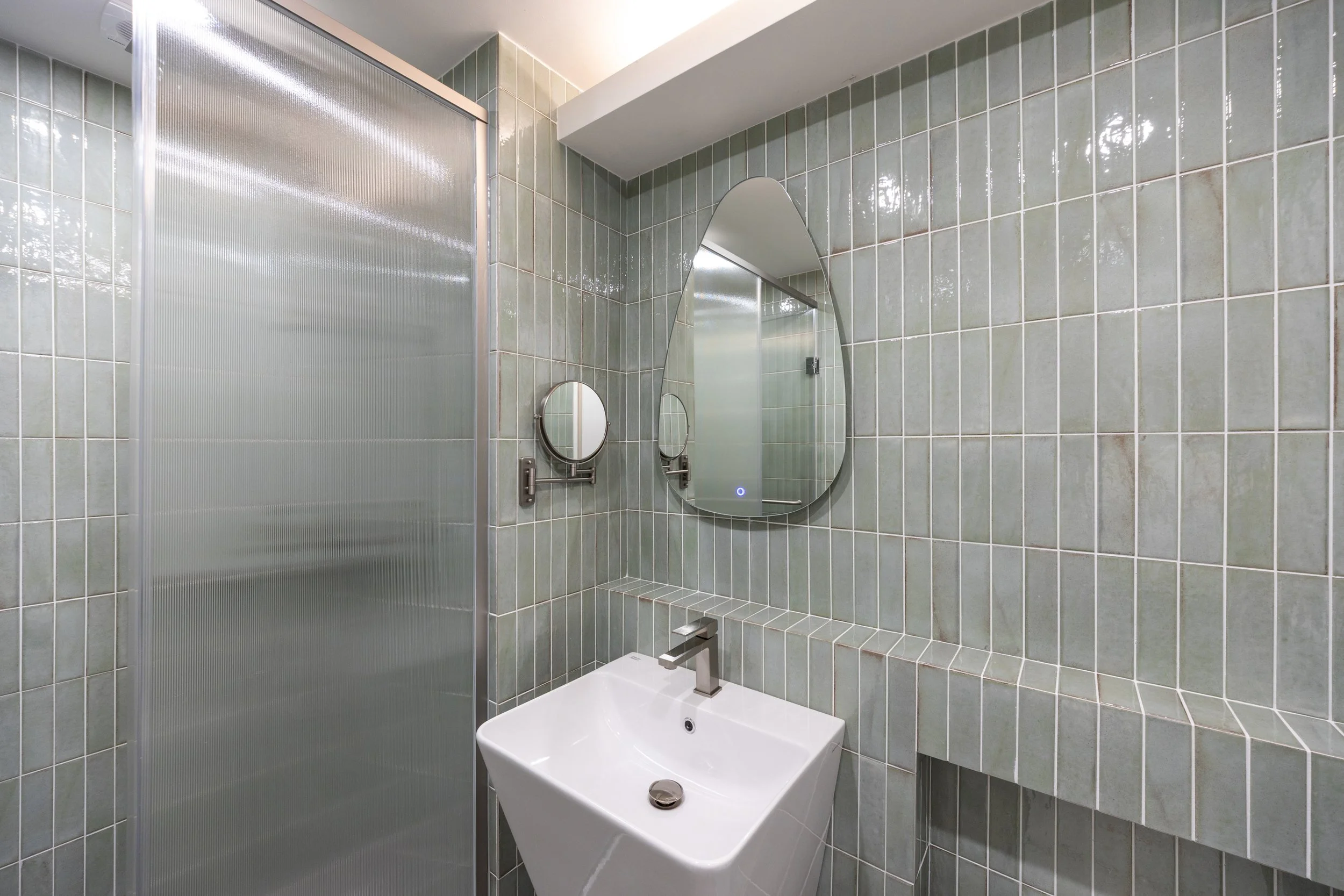 Modern bathroom with green tiles, a white sink, a large mirror, a circular magnifying mirror, and a frosted glass shower door.