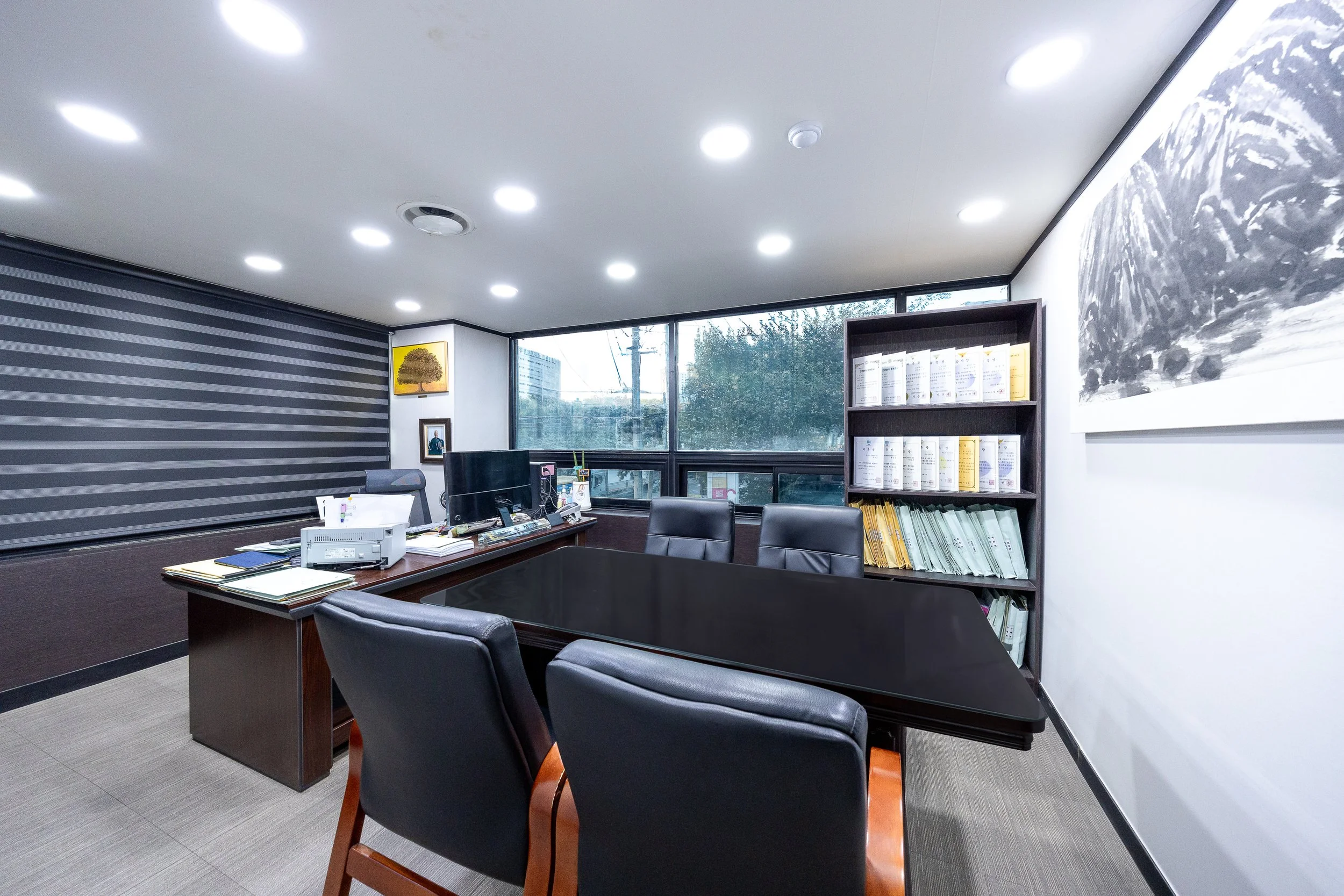 An office room with a large black conference table, several black chairs, a wooden desk with a computer and papers, a bookshelf with files, large windows, and artwork on the walls.