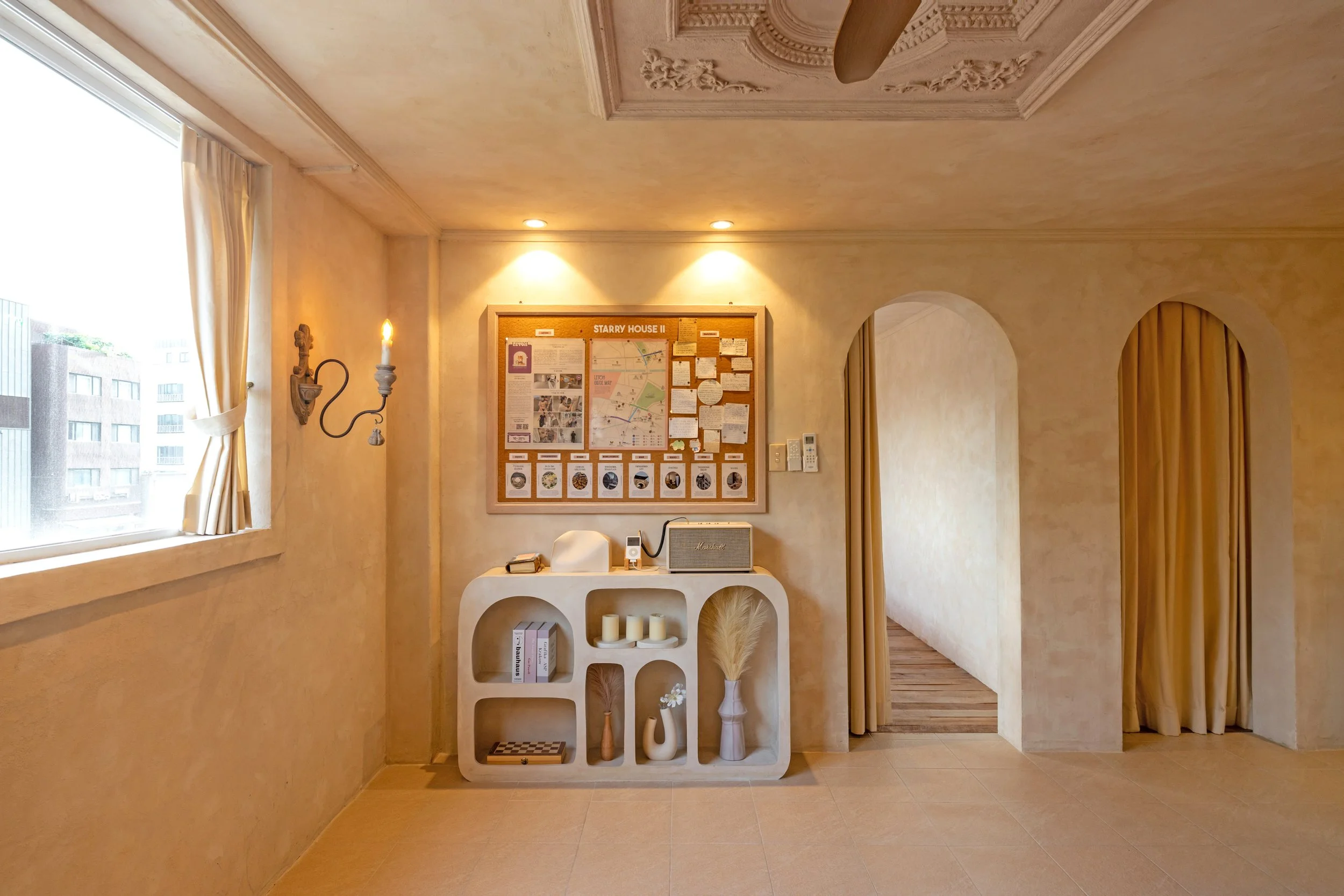 Interior of a cozy room with beige walls, a window with cream curtains, a wall-mounted candle sconce, a bulletin board titled 'Starry House II', and a white shelf with decorative items like candles, books, vases, and a mirror.