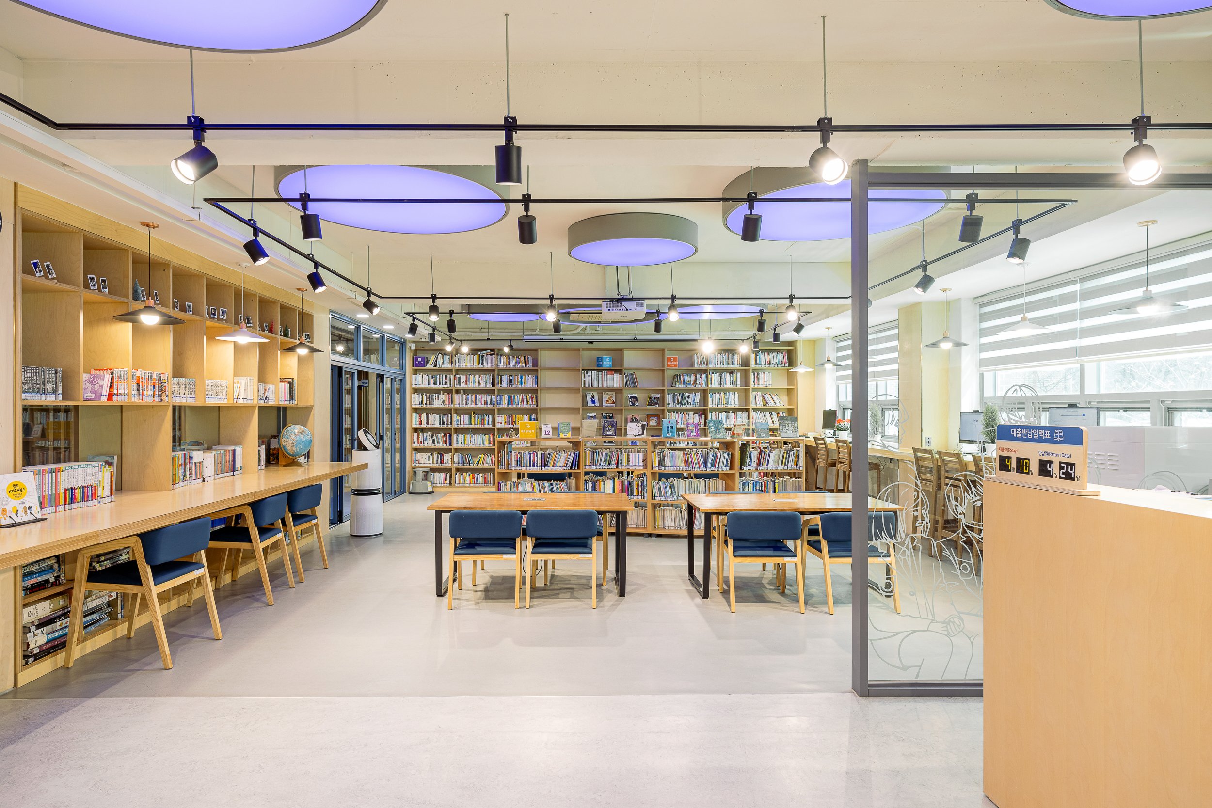 View of a modern library with bookshelves filled with books, a study table with chairs, and a reception desk. Large windows let in natural light, and ceiling lights illuminate the space.