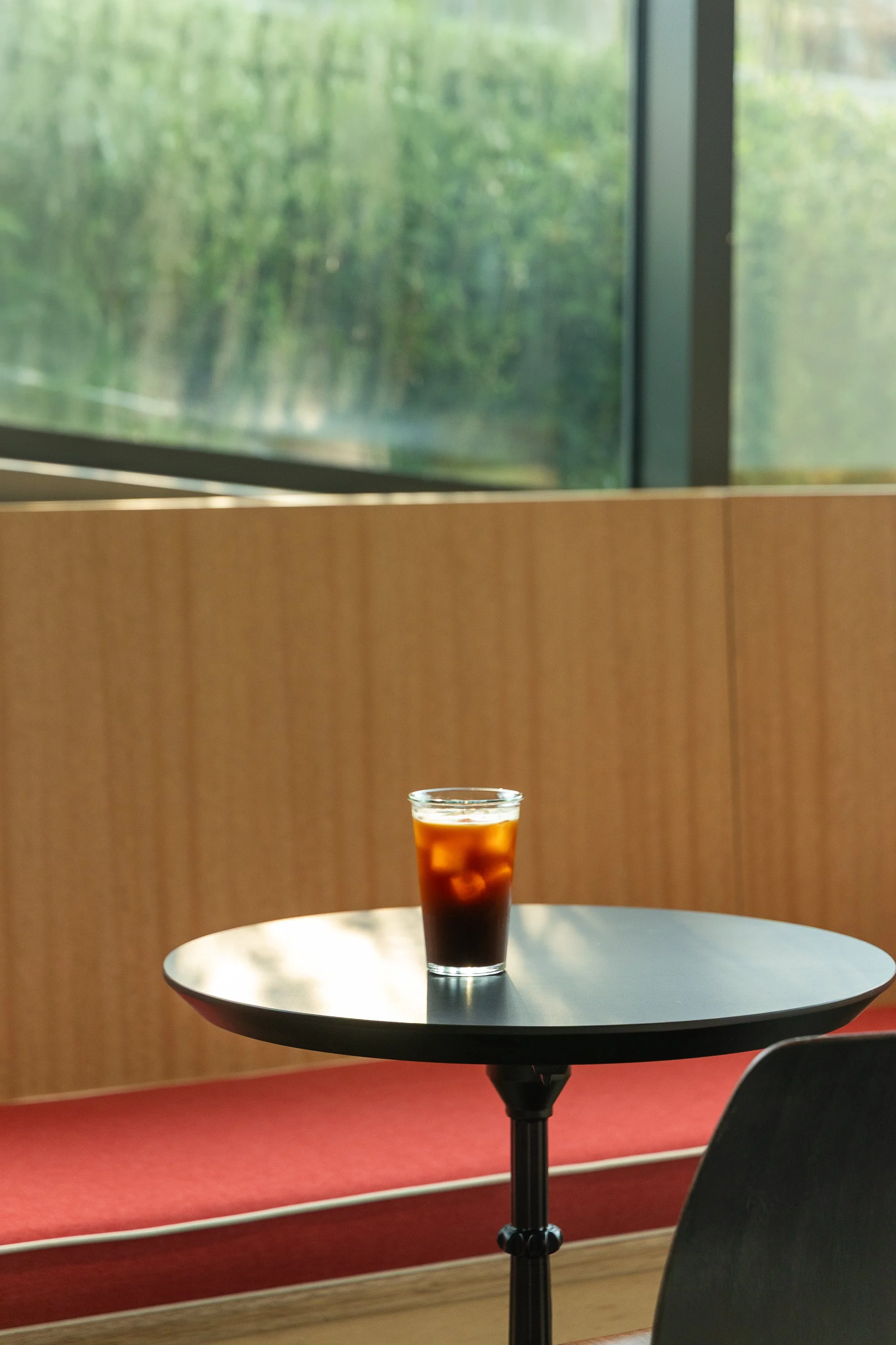 A glass of iced coffee on a round black table inside a cafe near a large window with a view of green trees.
