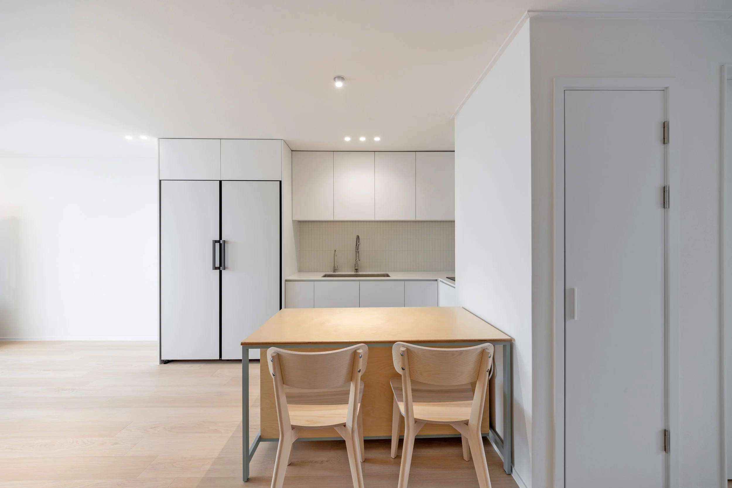 Minimalist kitchen with white cabinets, a wooden dining table, and two chairs, featuring a refrigerator, sink, and storage cabinets.