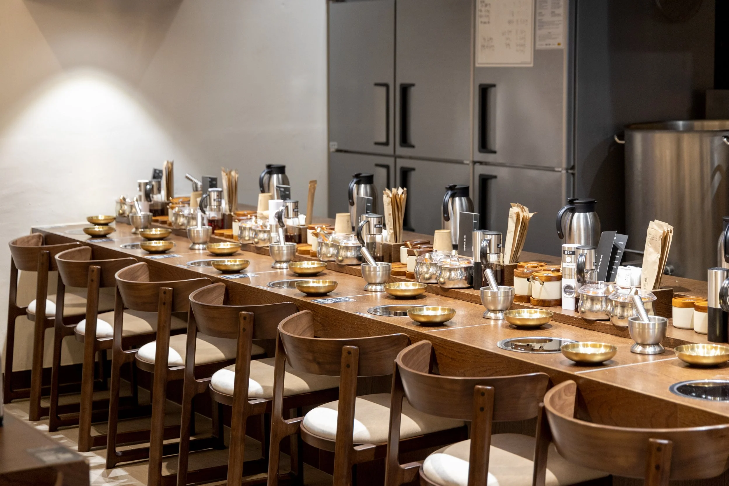 A restaurant or cafe counter with wooden chairs and a long wooden table set with metal bowls, cups, utensils, and coffee pots in front of a gray industrial-style kitchen.