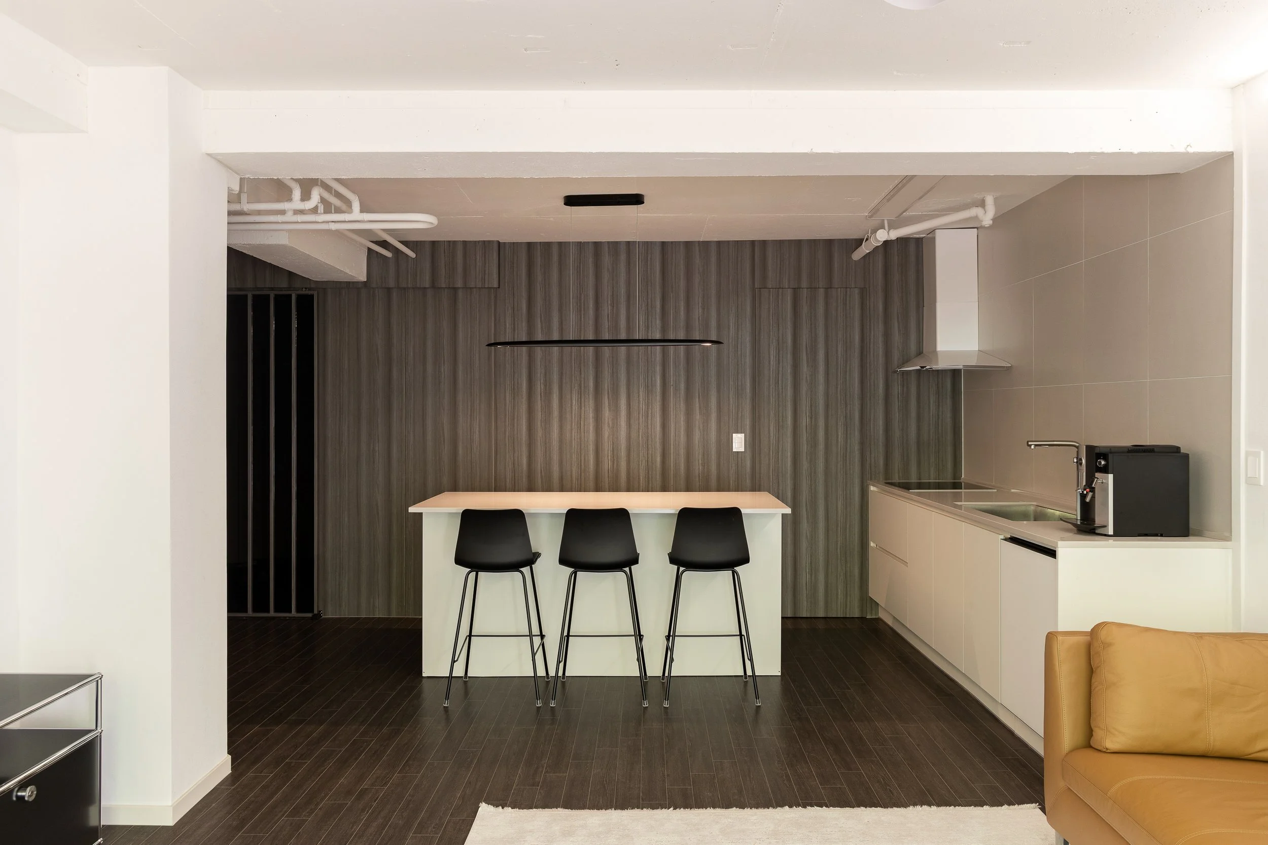 Modern kitchen with a white island, three black bar stools, dark wood floors, a gray textured wall, and a small kitchen area with a sink, coffee machine, and stainless steel range hood.