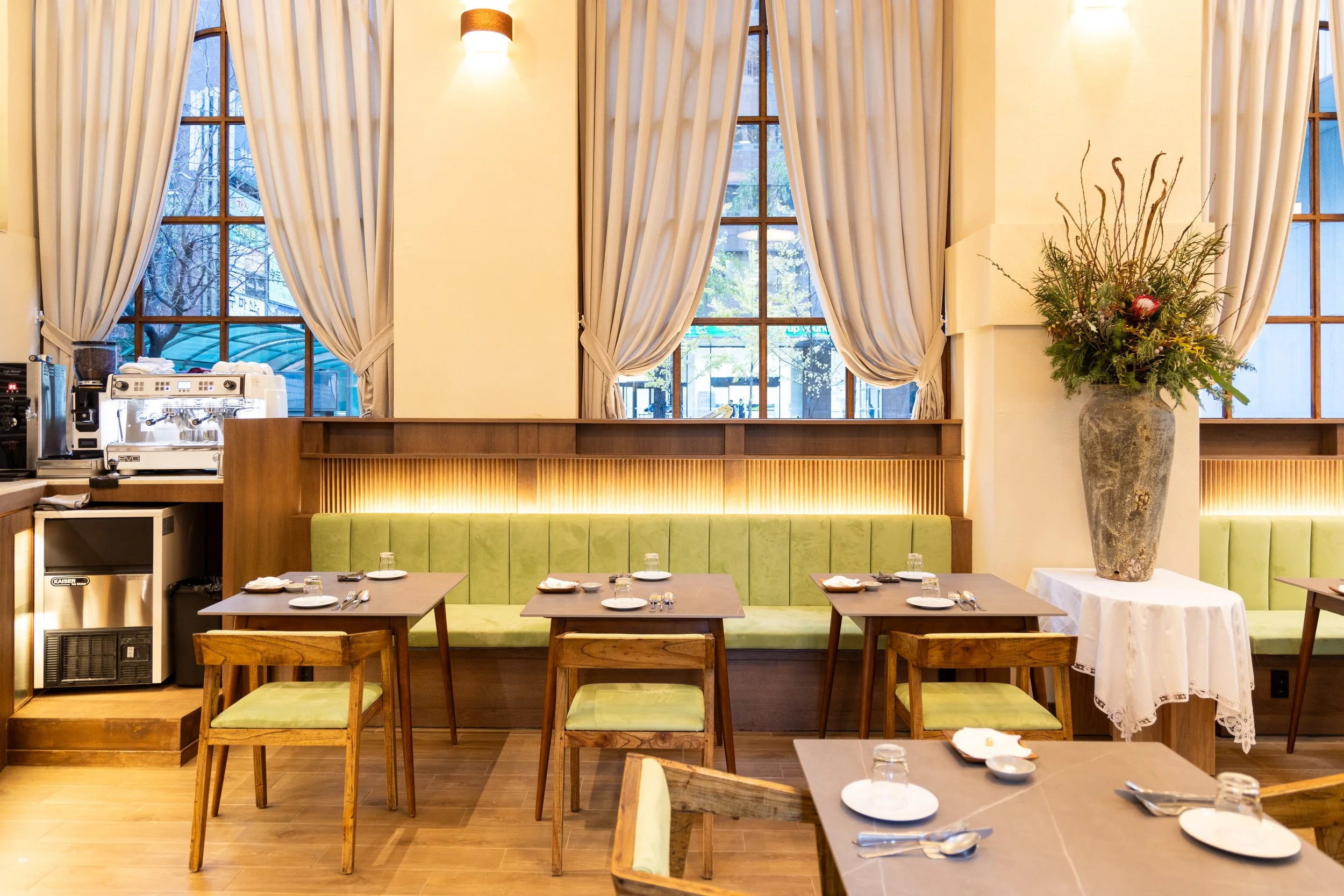 Interior of a restaurant with wooden tables and chairs, a green upholstered banquette, large windows with white curtains, and a tall vase with flowers on a table in the corner.