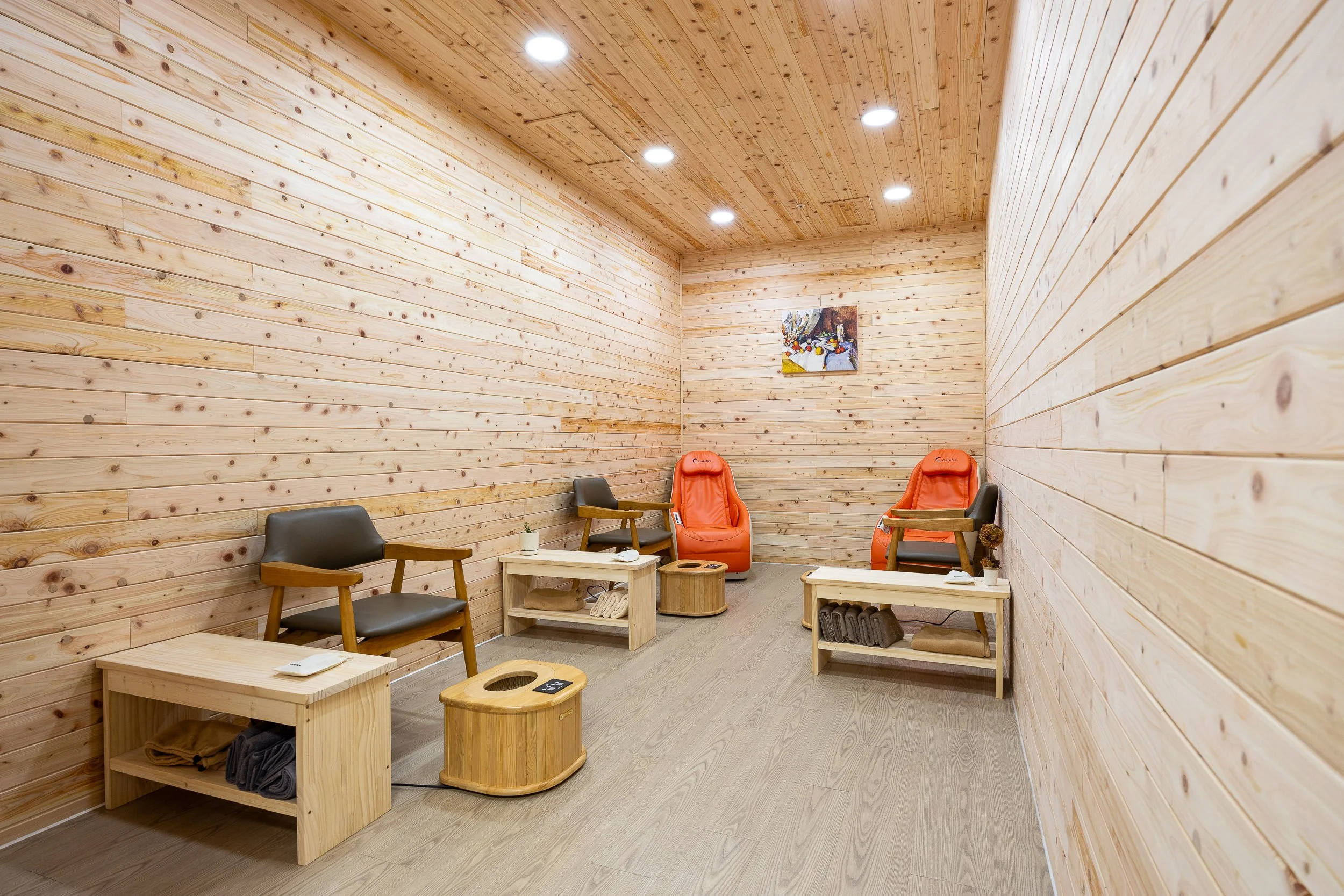 A serene room with wooden walls and ceiling, furnished with two black armchairs, two orange massage chairs, and small beige tables, some holding decorative items; a small painting hangs on the back wall.