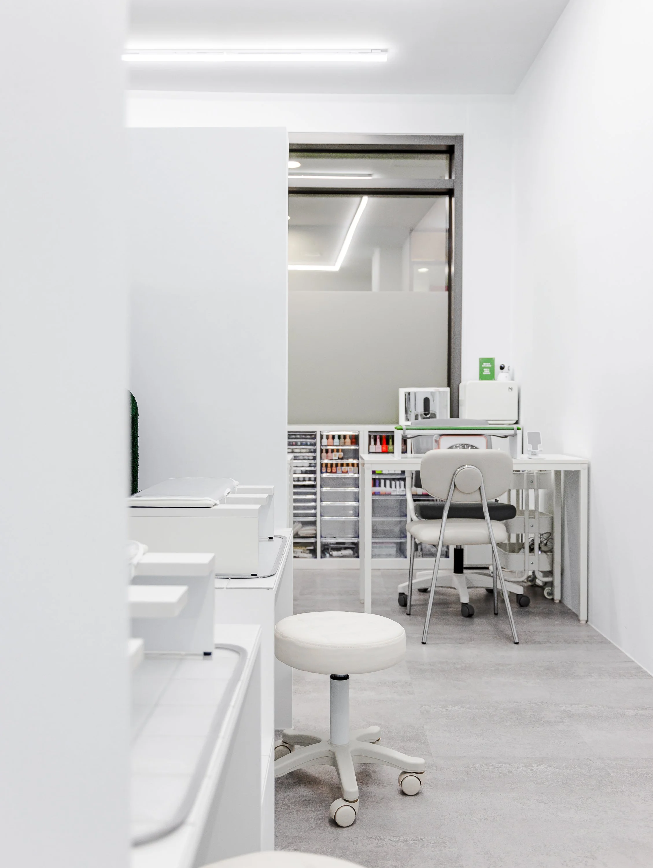 A clean, minimalist workspace with white walls, a white rolling chair, a desk with manicure supplies, and shelves with nail polish bottles in the background.